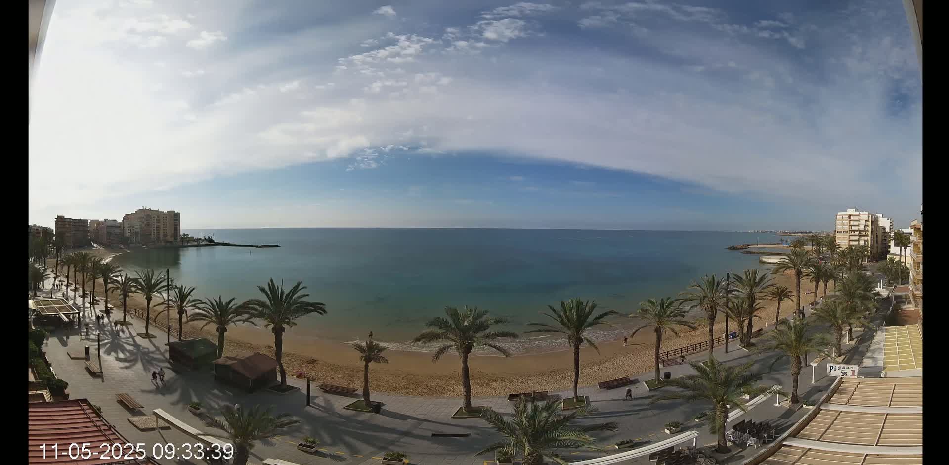 A sandy beach lined with palm trees and buildings is visible, under a partly cloudy sky, with calm blue water extending to the horizon.