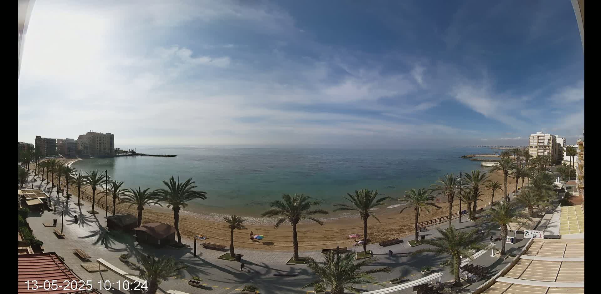 A sandy beach lined with palm trees and buildings is visible under a partly cloudy sky, with calm, turquoise water stretching to the horizon.