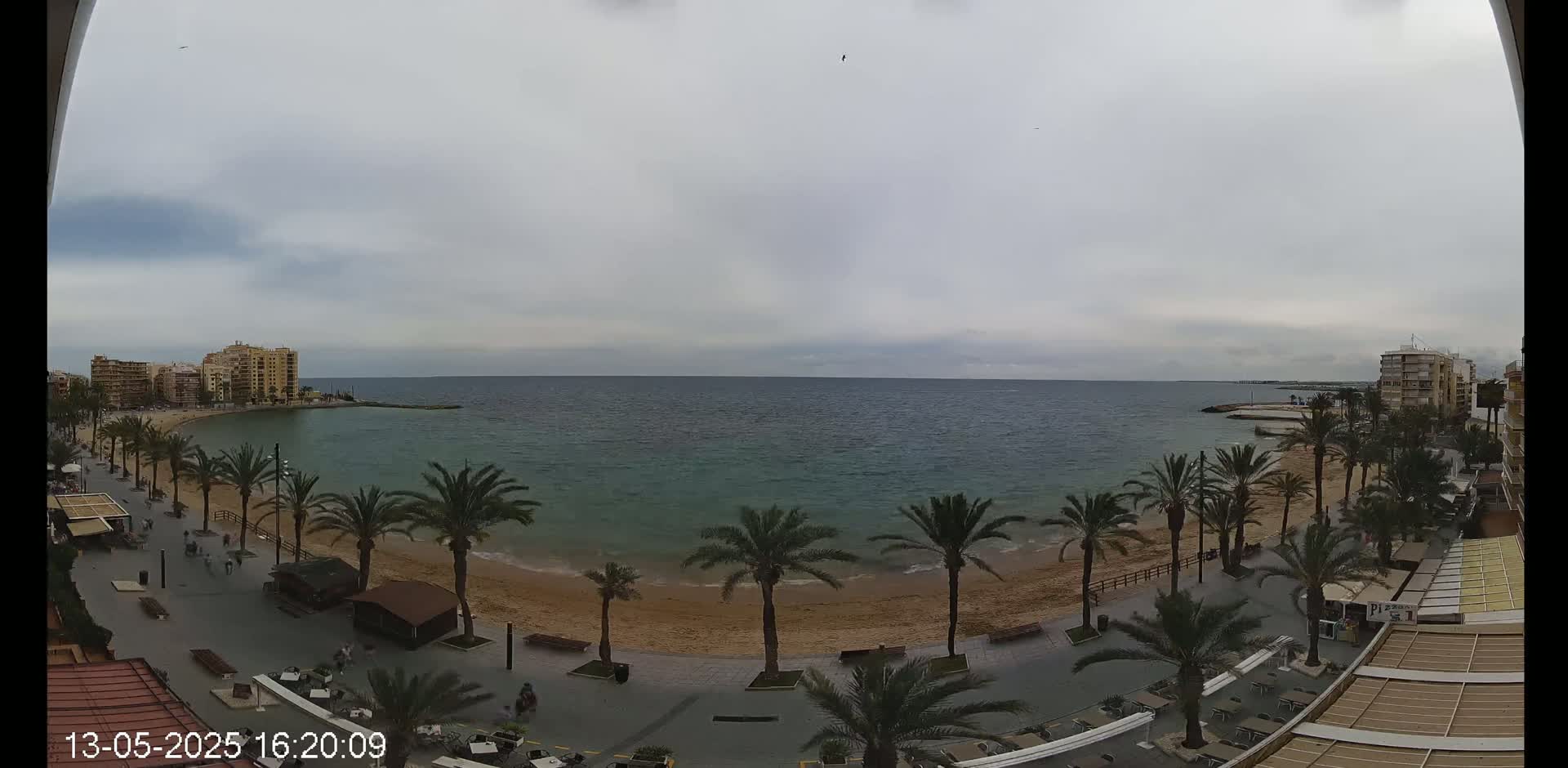 A mostly cloudy day overlooks a sandy beach lined with palm trees and buildings, next to a calm, teal sea.