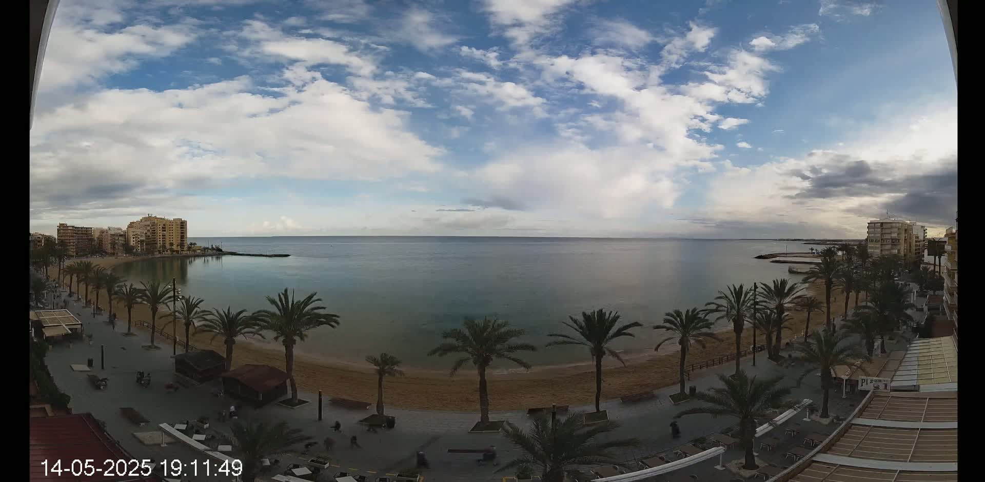 A partly cloudy day shows a sandy beach lined with palm trees and buildings overlooking a calm sea.