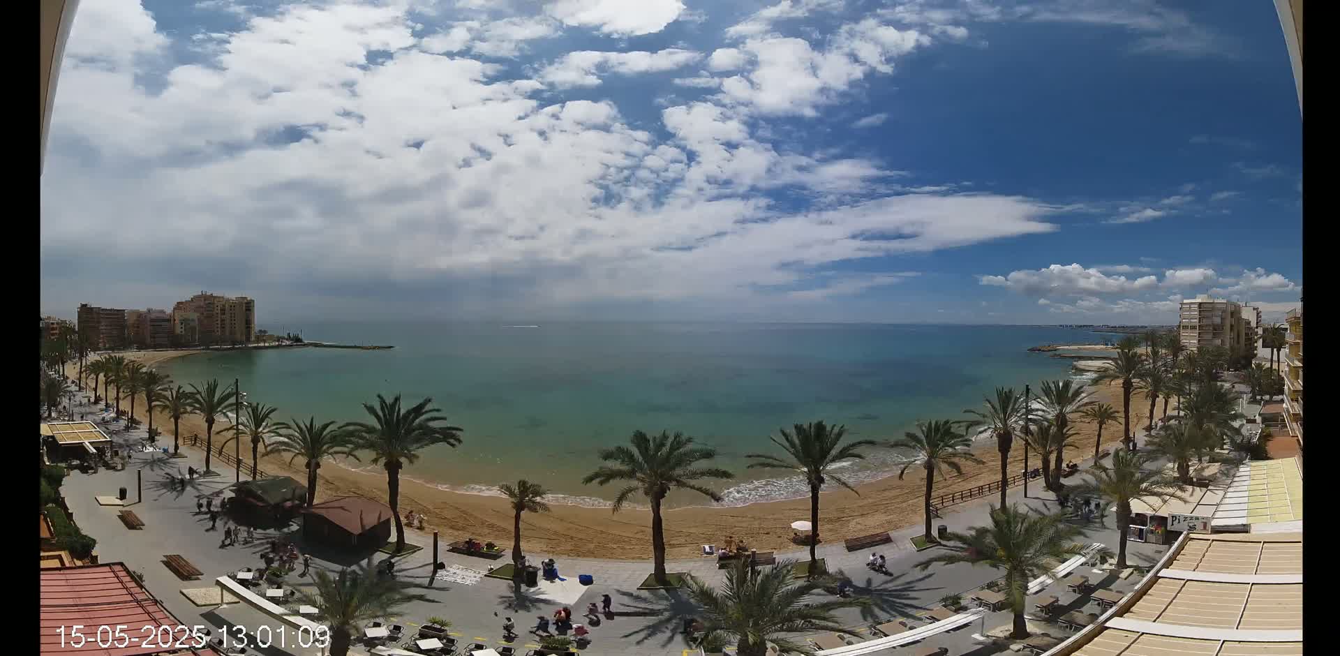 A partly cloudy sky overlooks a sandy beach lined with palm trees and buildings, a calm turquoise sea beyond.
