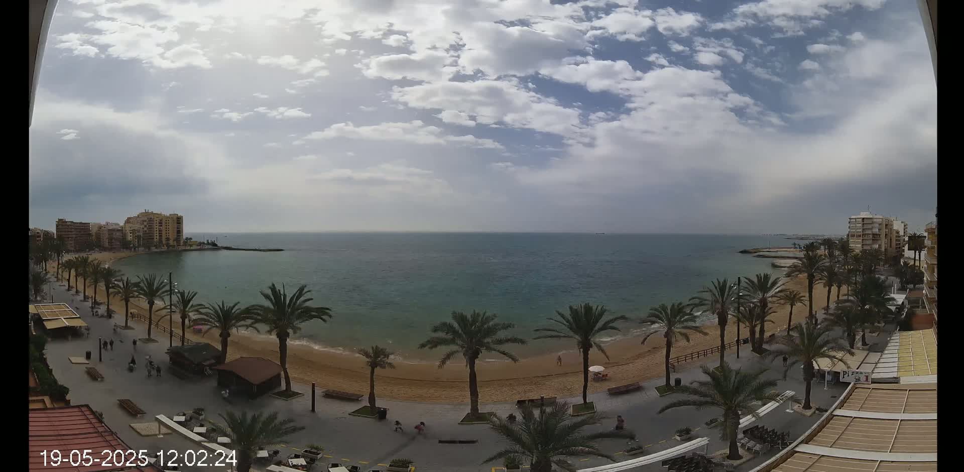 A partly cloudy day shows a sandy beach with palm trees lining a paved walkway next to calm blue ocean water and buildings in the distance.
