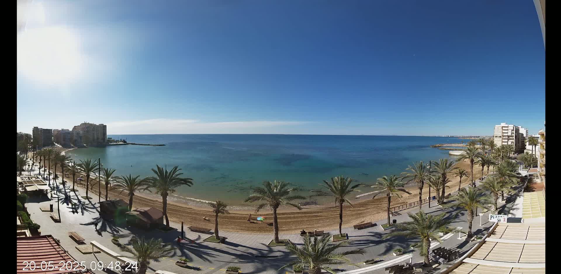 A sunny day reveals a sandy beach lined with palm trees and bordered by buildings, overlooking a calm, clear turquoise sea.