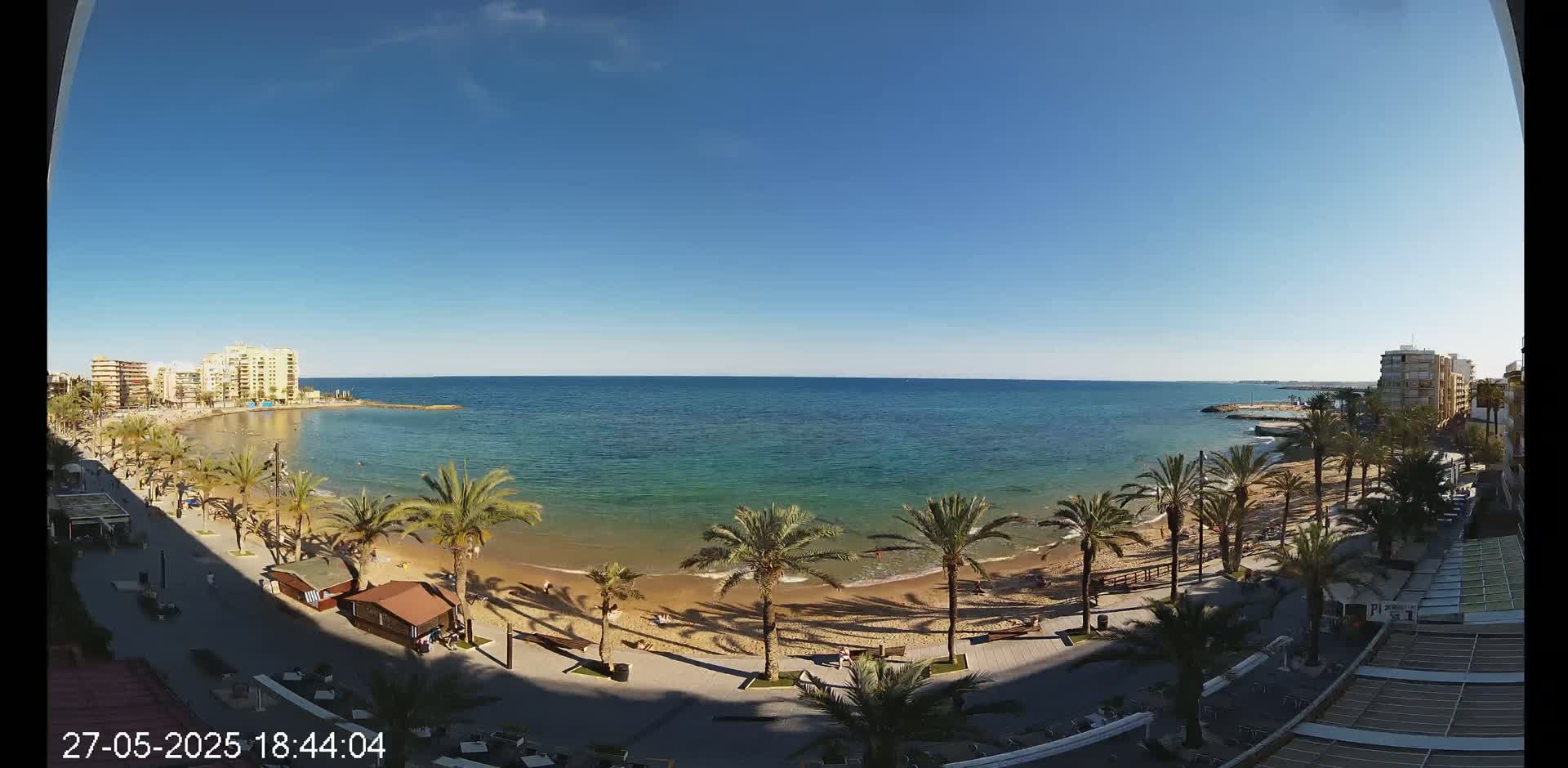A sunny day reveals a sandy beach lined with palm trees, buildings, and a calm, turquoise ocean.