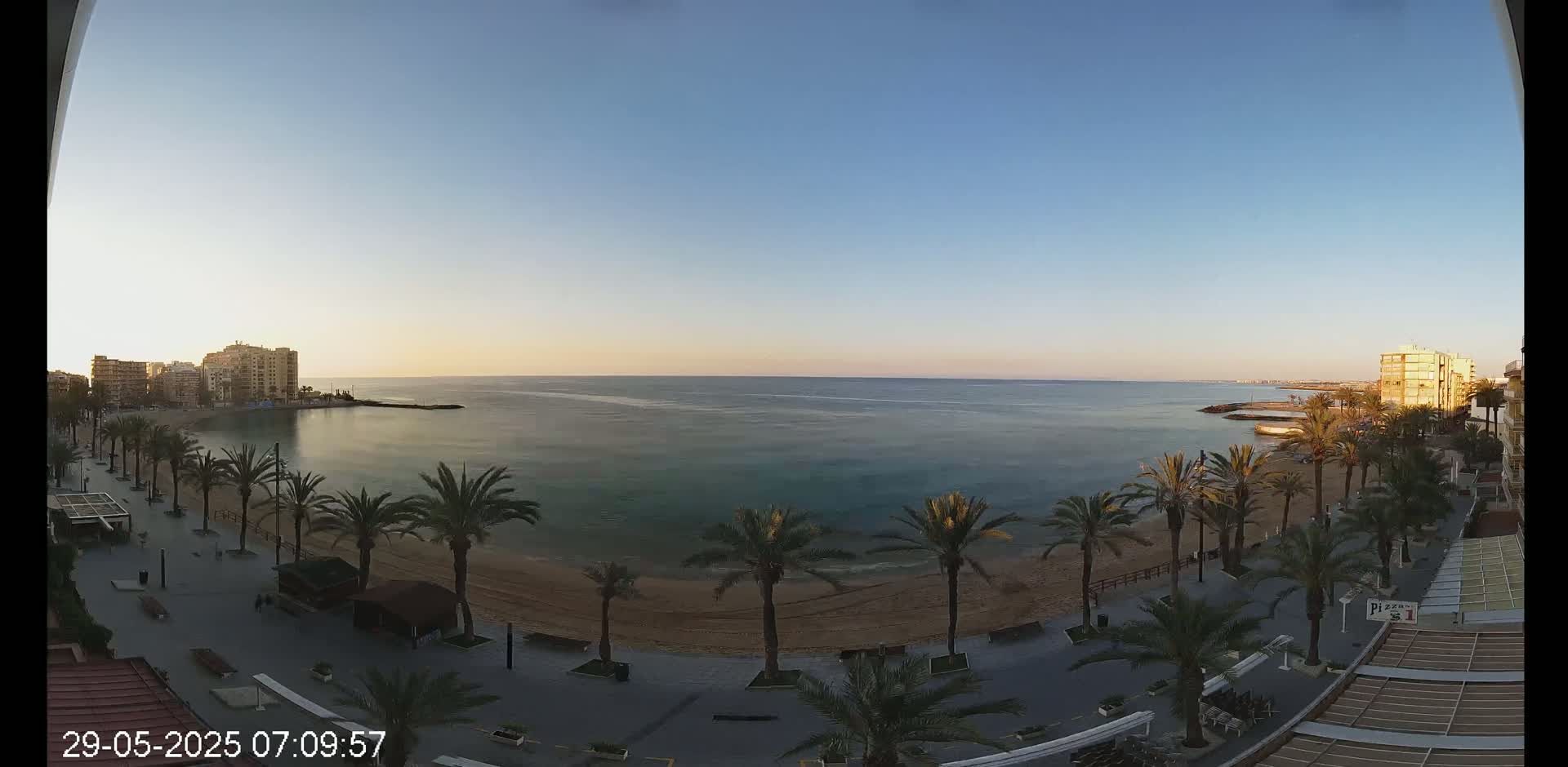 A calm, clear morning reveals a sandy beach lined with palm trees, a boardwalk, and buildings along a tranquil body of water.