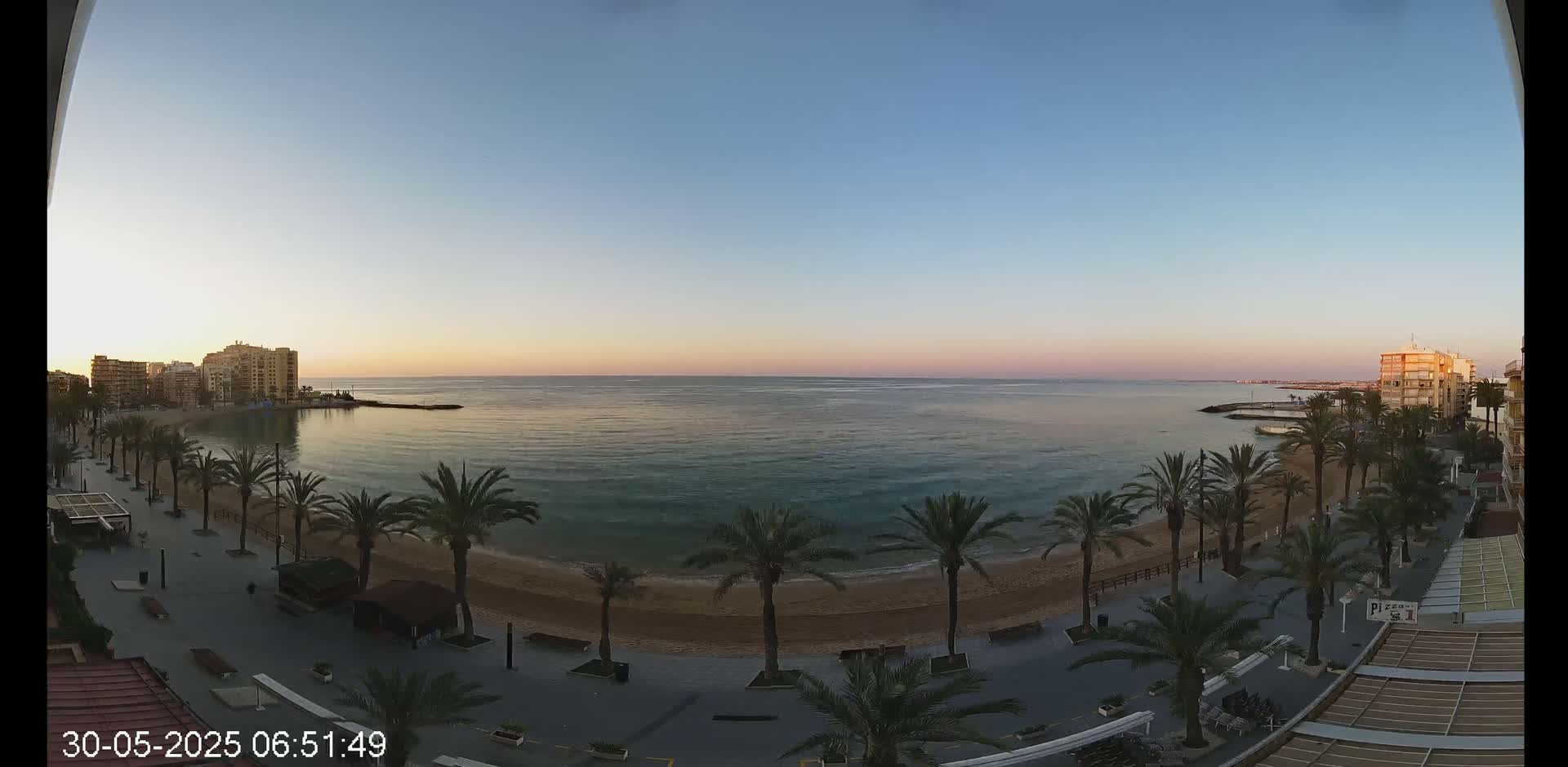 A calm, twilight sea laps a sandy beach lined with palm trees and beachfront buildings under a clear, pale sky.