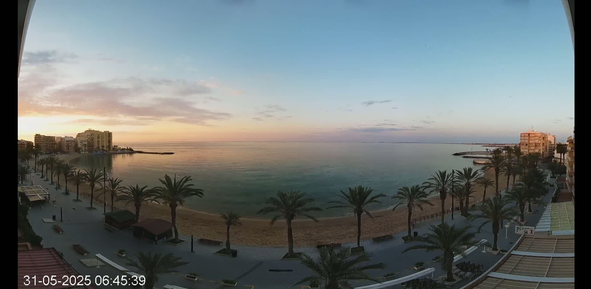 A tranquil sunrise illuminates a sandy beach lined with palm trees and buildings, overlooking a calm sea.