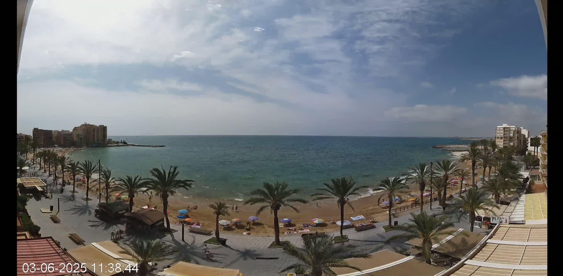 A partly sunny day at a beach, showing a sandy shore lined with palm trees, umbrellas, and people, with buildings visible in the background.