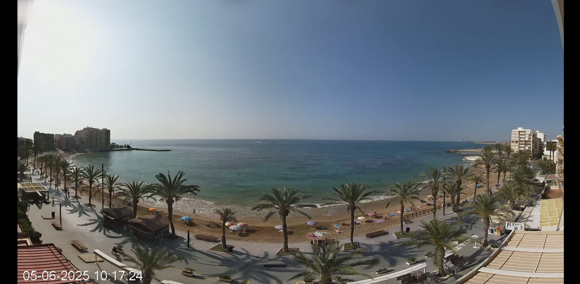 A sunny day at a sandy beach, lined with palm trees and bordered by buildings, shows a calm ocean with a few people scattered on the shore.