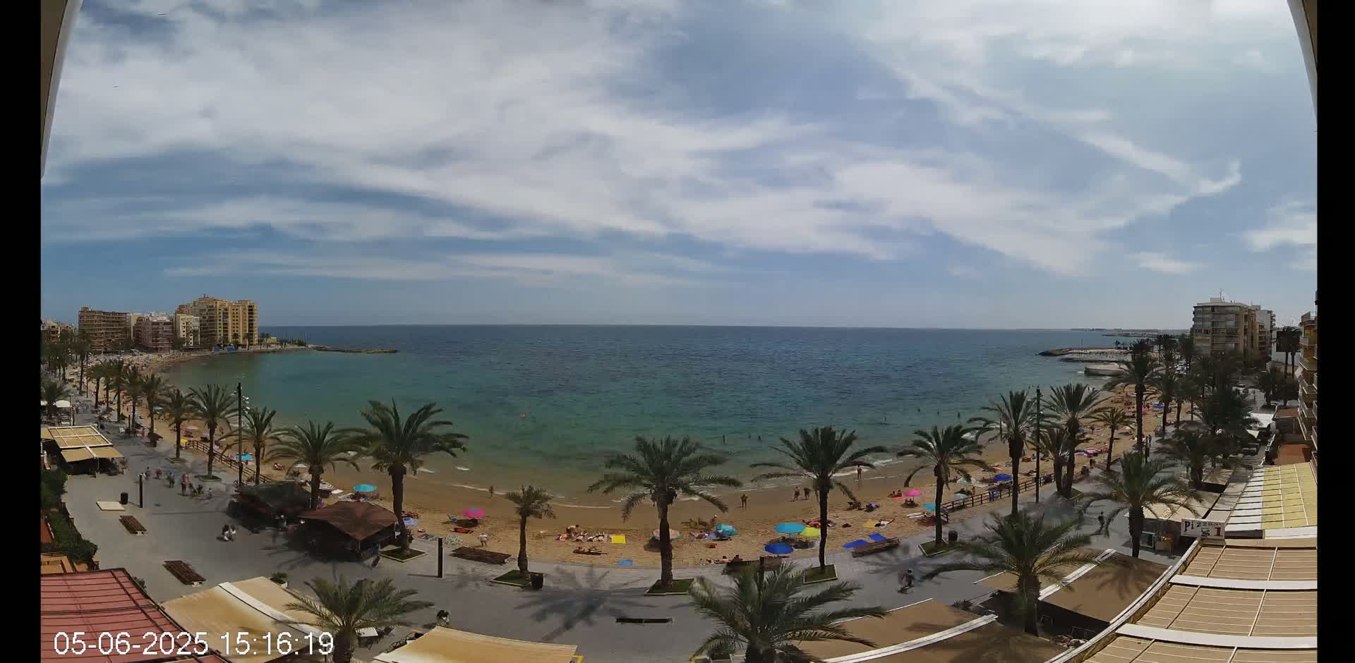 A partly cloudy day shows a sandy beach with colorful umbrellas, palm trees, and people enjoying the sun along a calm, teal-colored sea, bordered by buildings and a promenade.