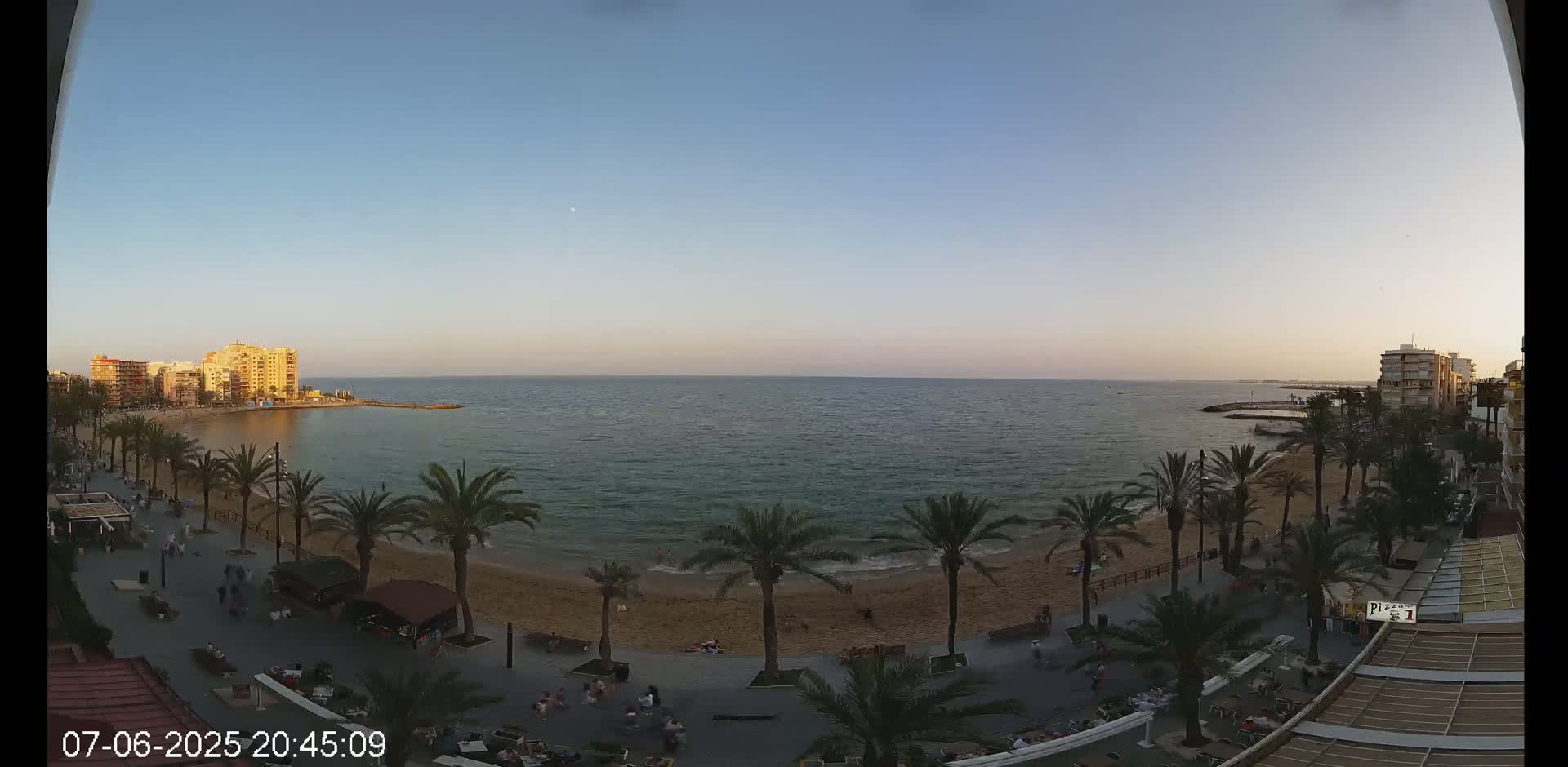 A sandy beach with palm trees and buildings is visible at sunset, under a clear sky, next to a calm sea.
