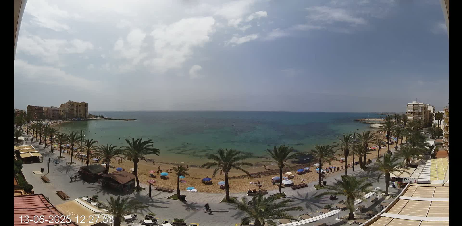 A partly cloudy day shows a sandy beach with palm trees and colorful umbrellas, a boardwalk with people walking, and buildings along a calm, teal-colored sea.