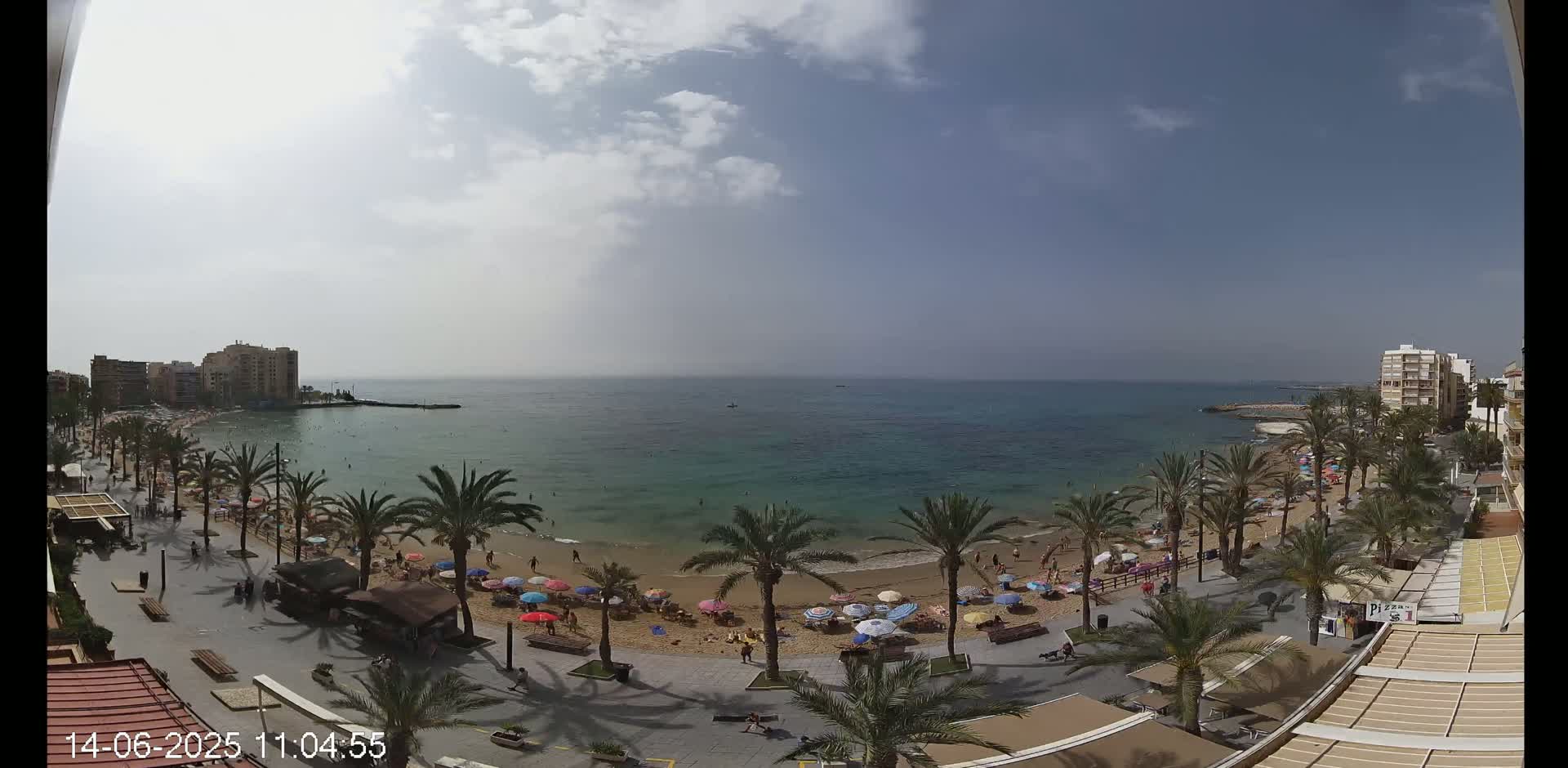 A partly cloudy day shows a beach with people, umbrellas, and palm trees lining a promenade next to a calm, turquoise ocean.