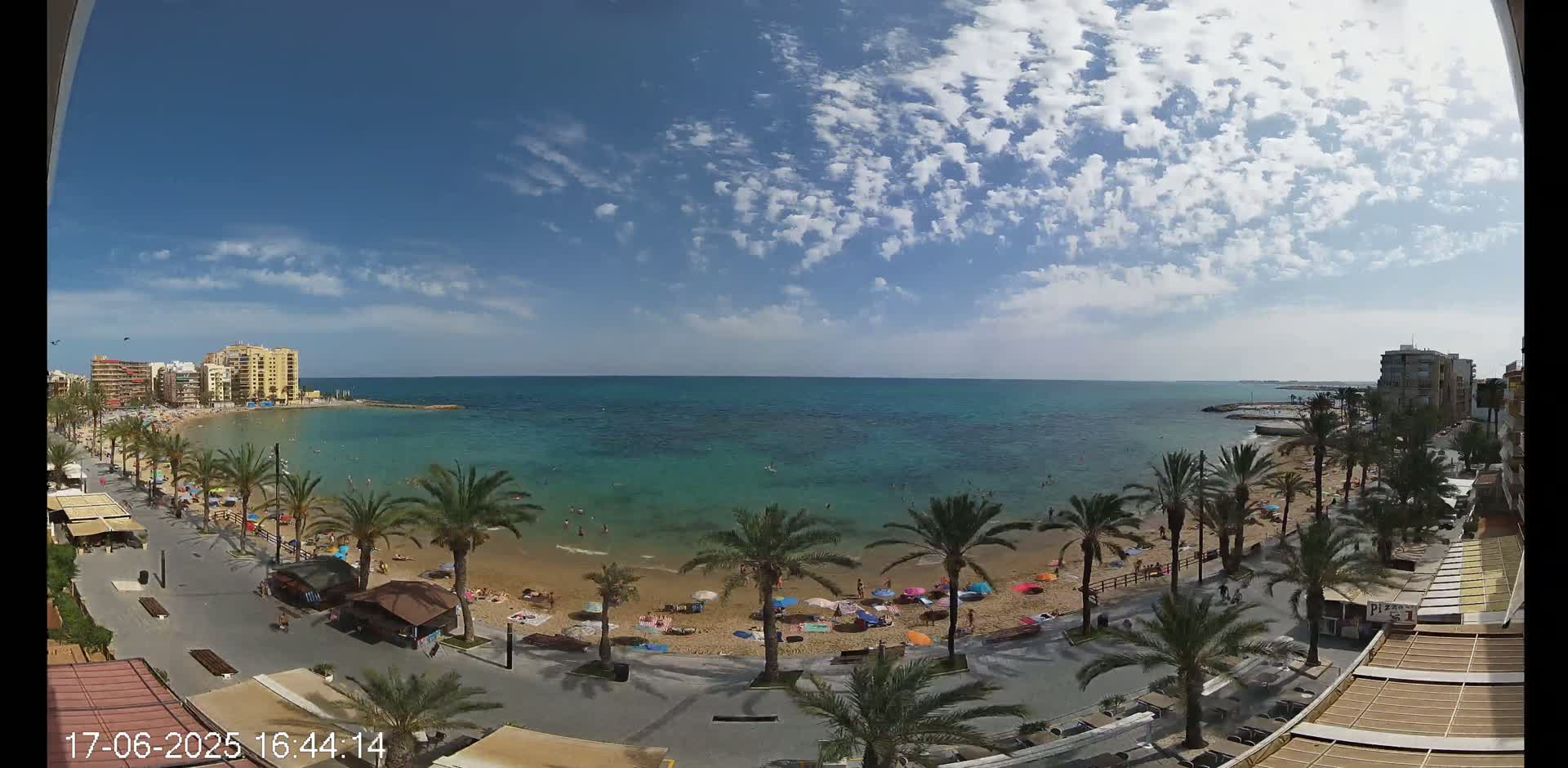A sunny day at a beach with a mostly clear sky and scattered clouds shows a sandy beach with people swimming and relaxing under umbrellas, palm trees lining a boardwalk, and buildings in the background.