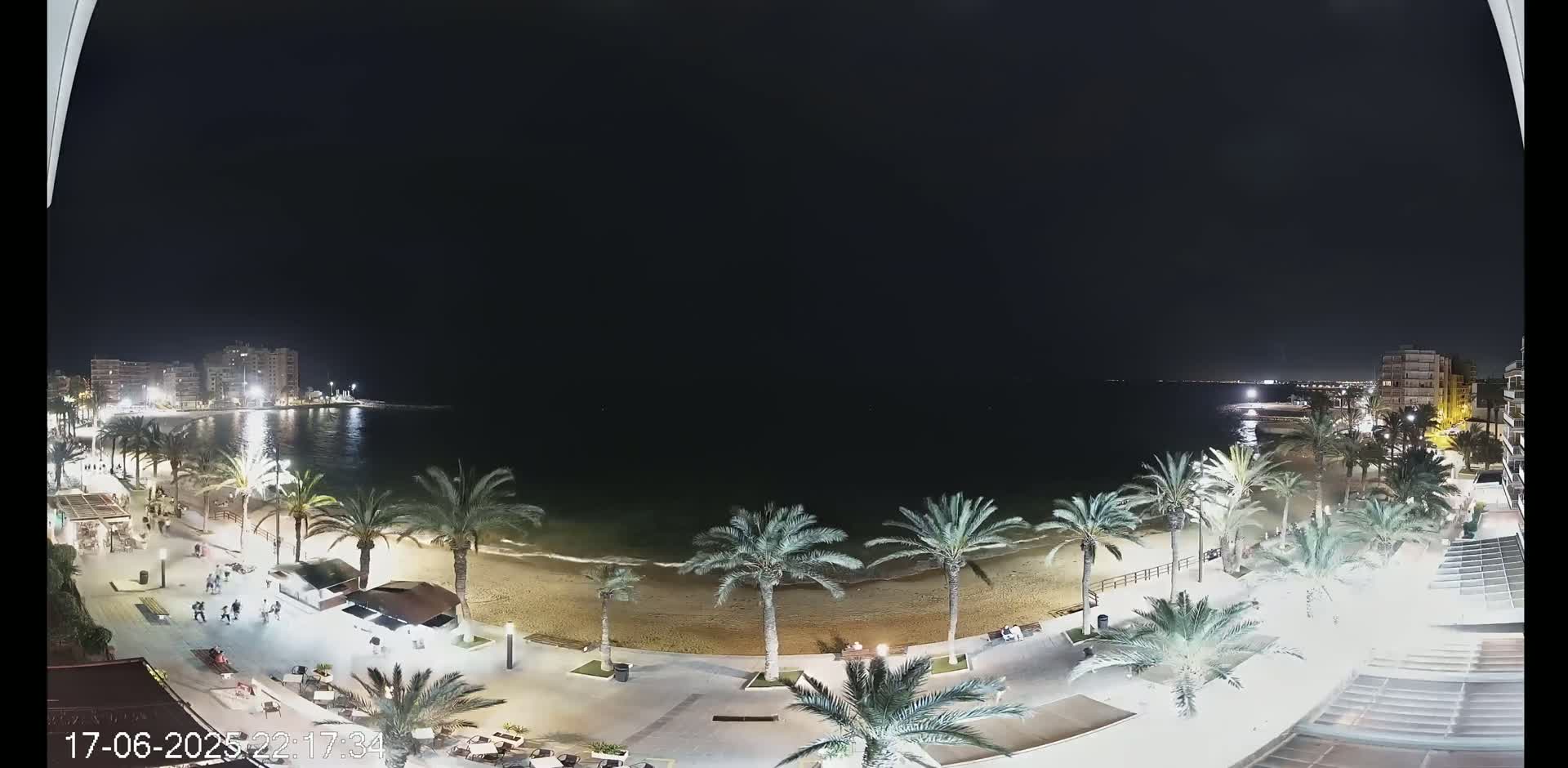 A nighttime view of a beach with palm trees, buildings, and people, under a clear dark sky.