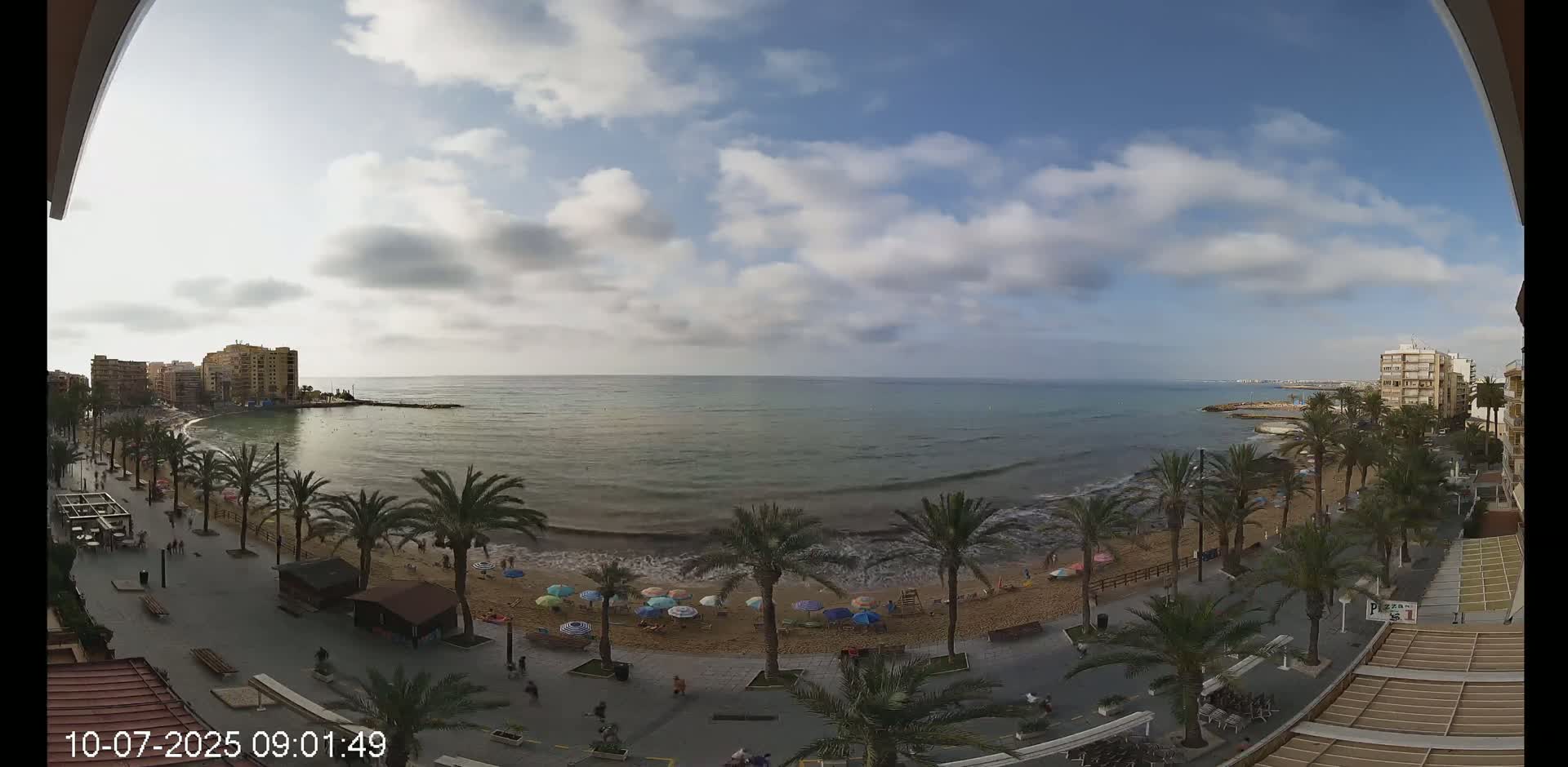 A partly cloudy day at a beachside promenade, with palm trees lining the sandy beach and buildings visible in the distance.