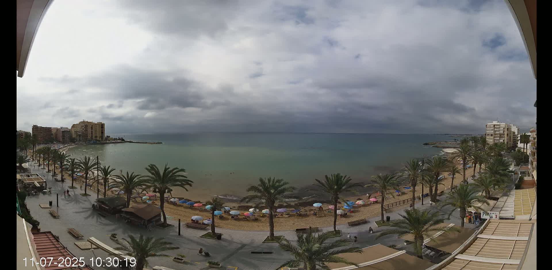A mostly cloudy day overlooks a sandy beach with palm trees and colorful umbrellas, a calm sea, and buildings in the distance.