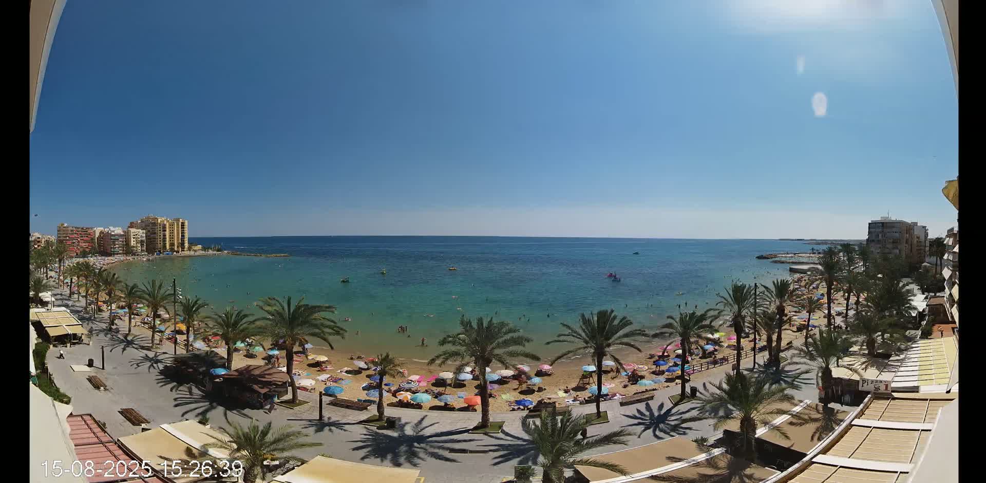 A sunny day shows a beach with colorful umbrellas and palm trees lining a boardwalk,  buildings in the background and calm blue water beyond.