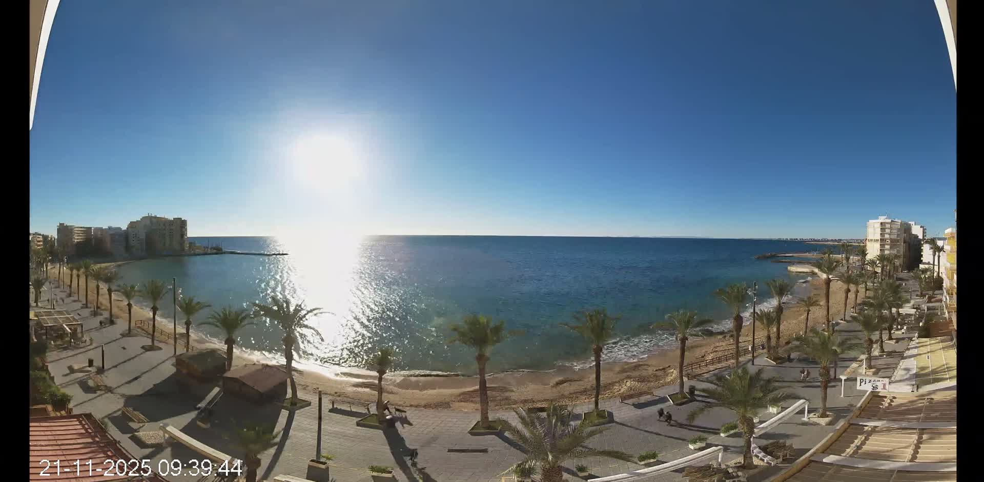 A wide panoramic view captures a sunny coastal bay with a sandy beach, a palm-lined promenade, and buildings along the shore, under a clear blue sky on a bright day.