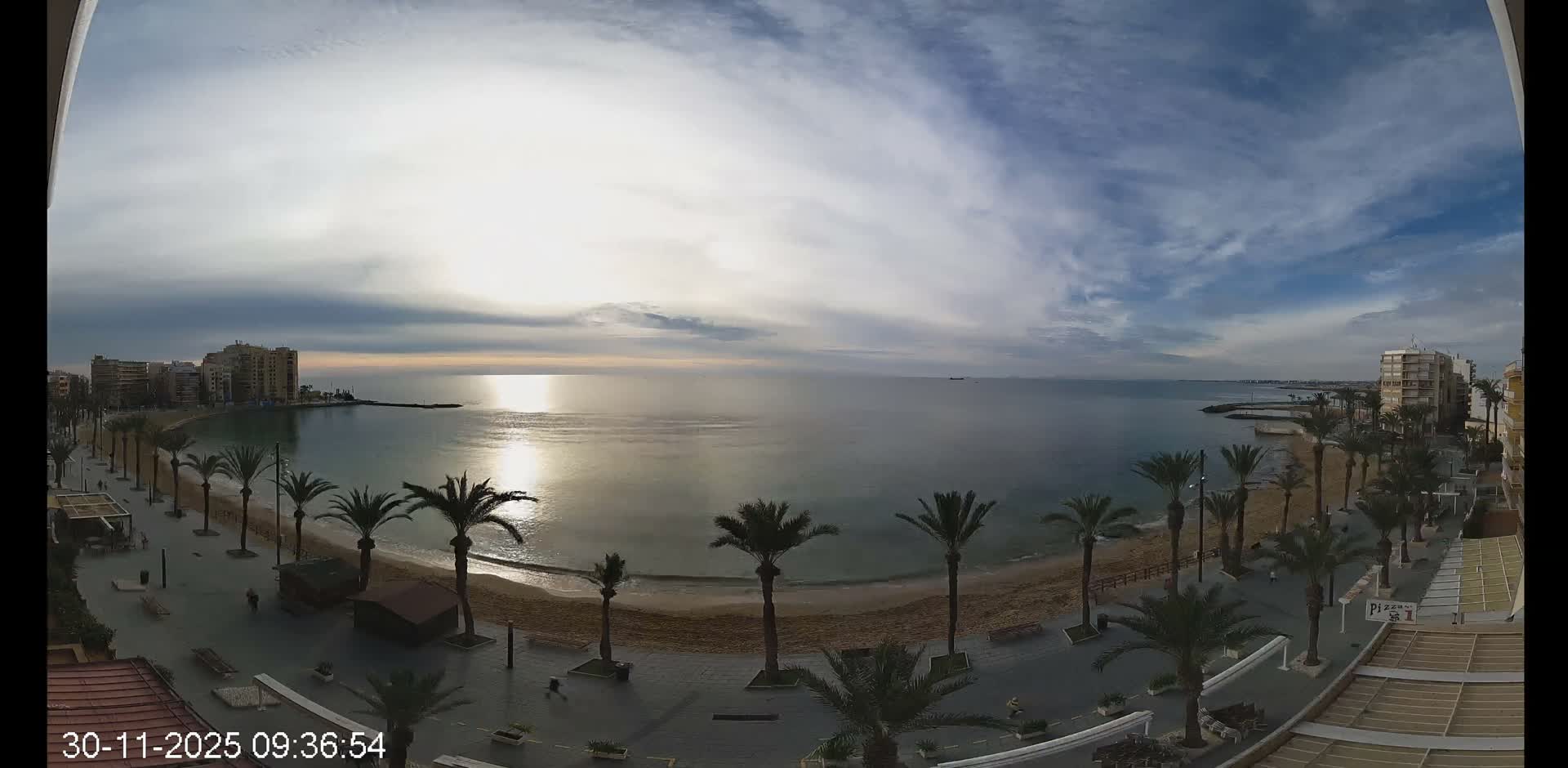 A wide panoramic view captures a tranquil coastal bay with brilliant sunlight reflecting on the water, a sandy beach, and a palm-lined promenade backed by buildings, all under a partly cloudy morning sky.