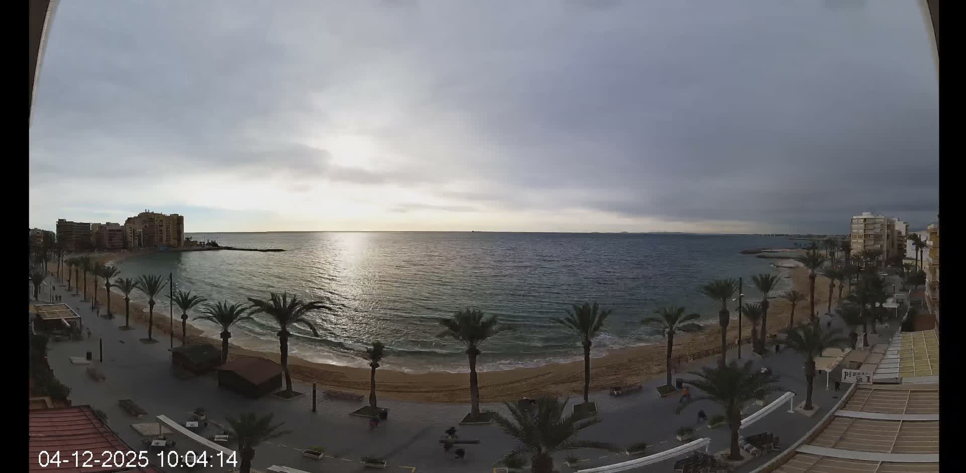 An elevated panoramic view captures a curving sandy beach and a palm-lined promenade bordering a coastal town, with sunlight reflecting on the gentle waves of the sea beneath an overcast sky.