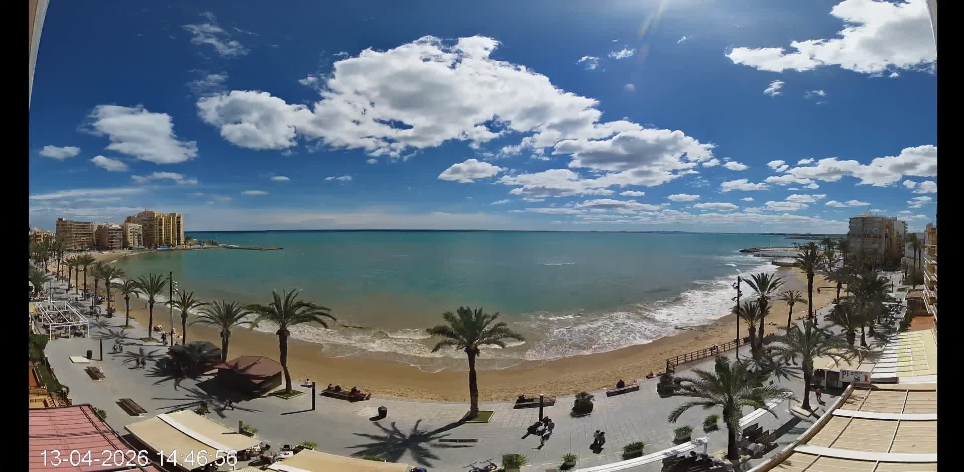 An elevated panoramic view captures a curving sandy beach and a palm-lined promenade bordering a coastal town, with sunlight reflecting on the gentle waves of the sea beneath an overcast sky.