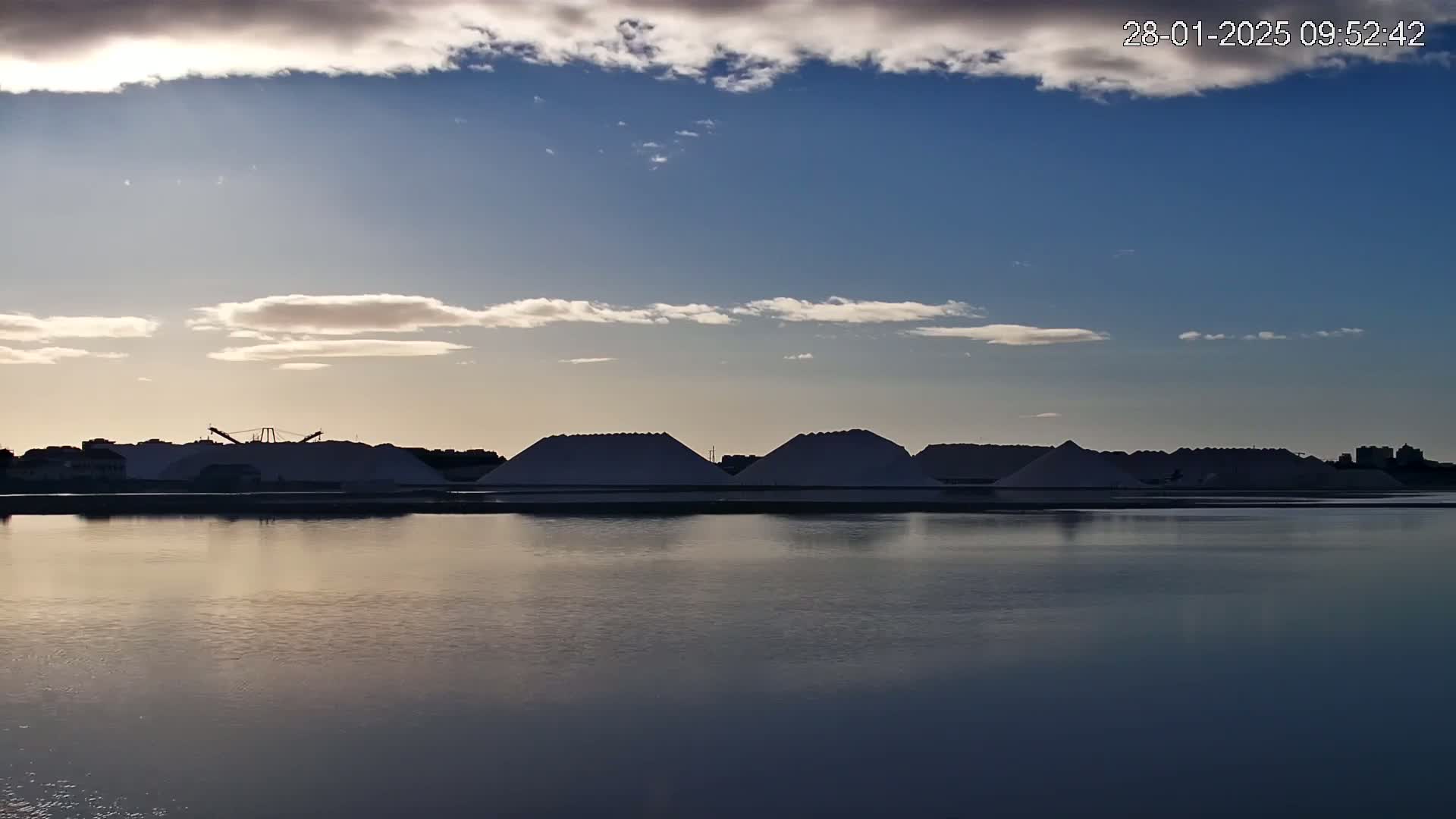 Several large piles of what appears to be salt are situated near a calm body of water under a partly cloudy sky.