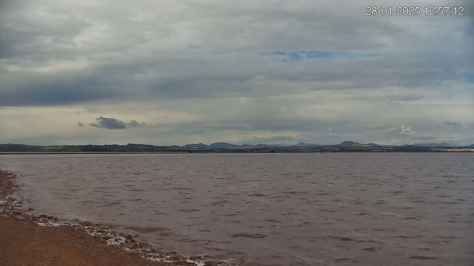 A slightly choppy, pinkish-brown body of water stretches to a distant shoreline of low hills under a cloudy sky.