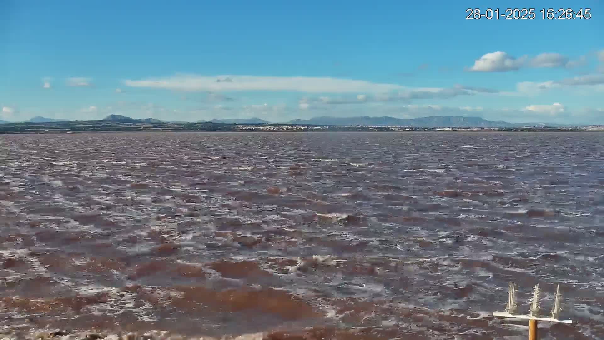 A choppy, reddish-brown body of water stretches towards a distant mountain range under a mostly clear, blue sky with a few clouds.