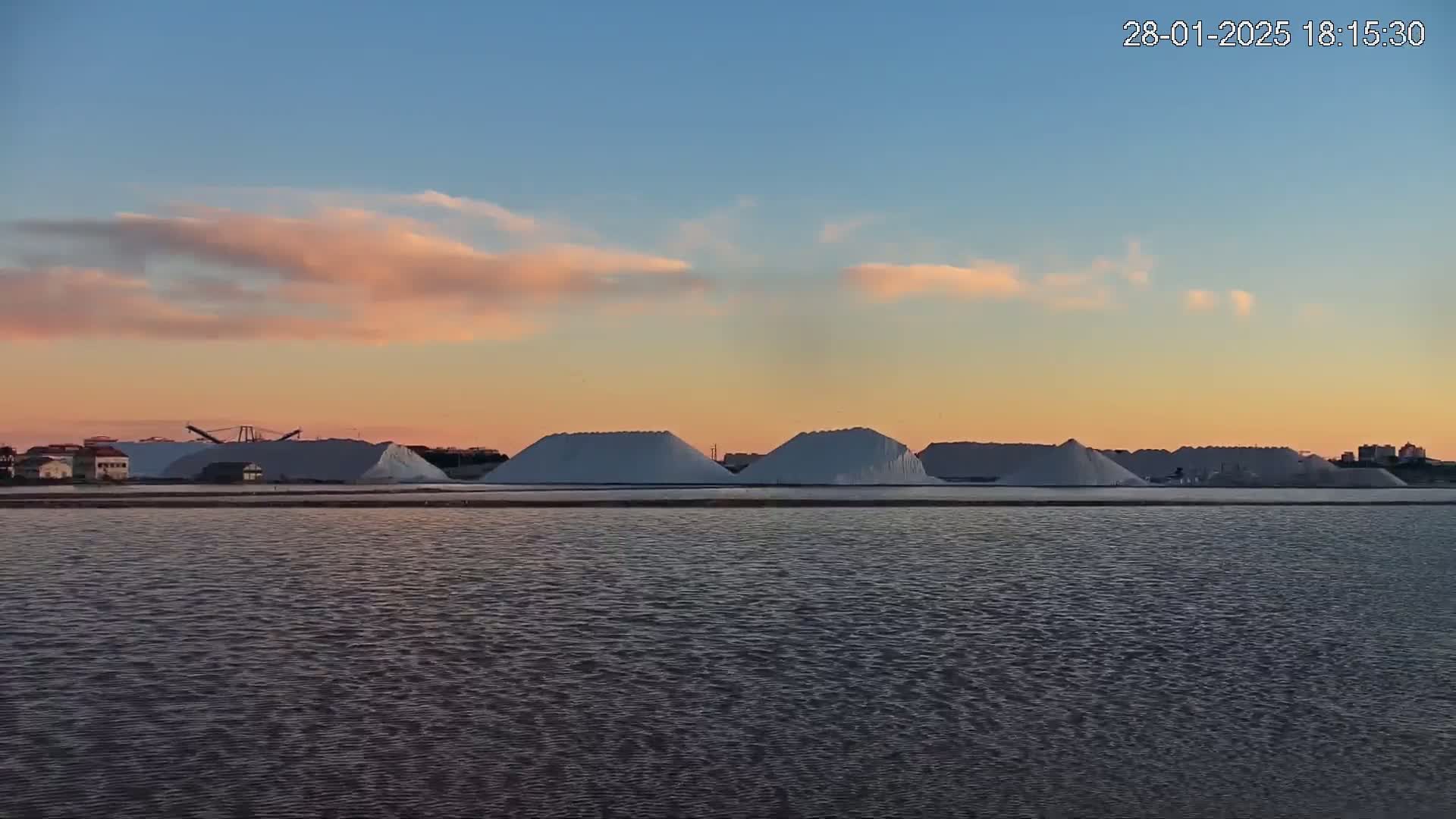 Several large piles of salt are situated on the far shore of a calm body of water under a sunset sky with soft pink clouds.