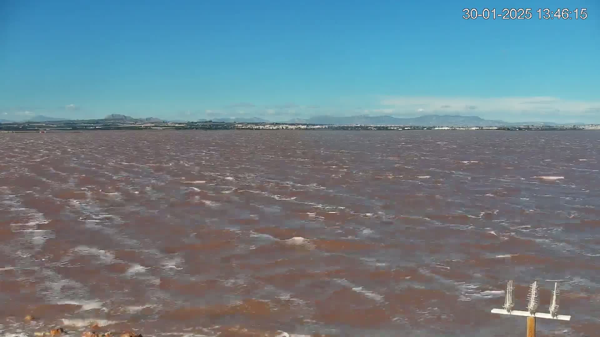 A reddish-brown body of water with small waves under a clear blue sky shows a distant shoreline with mountains in the background.