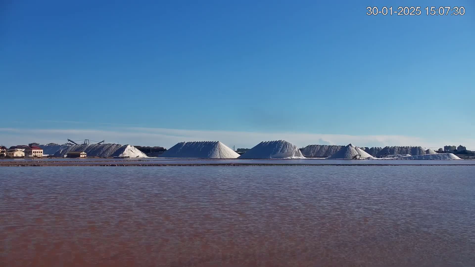 Under a clear blue sky, several large piles of white salt are situated near a body of reddish-pink water.