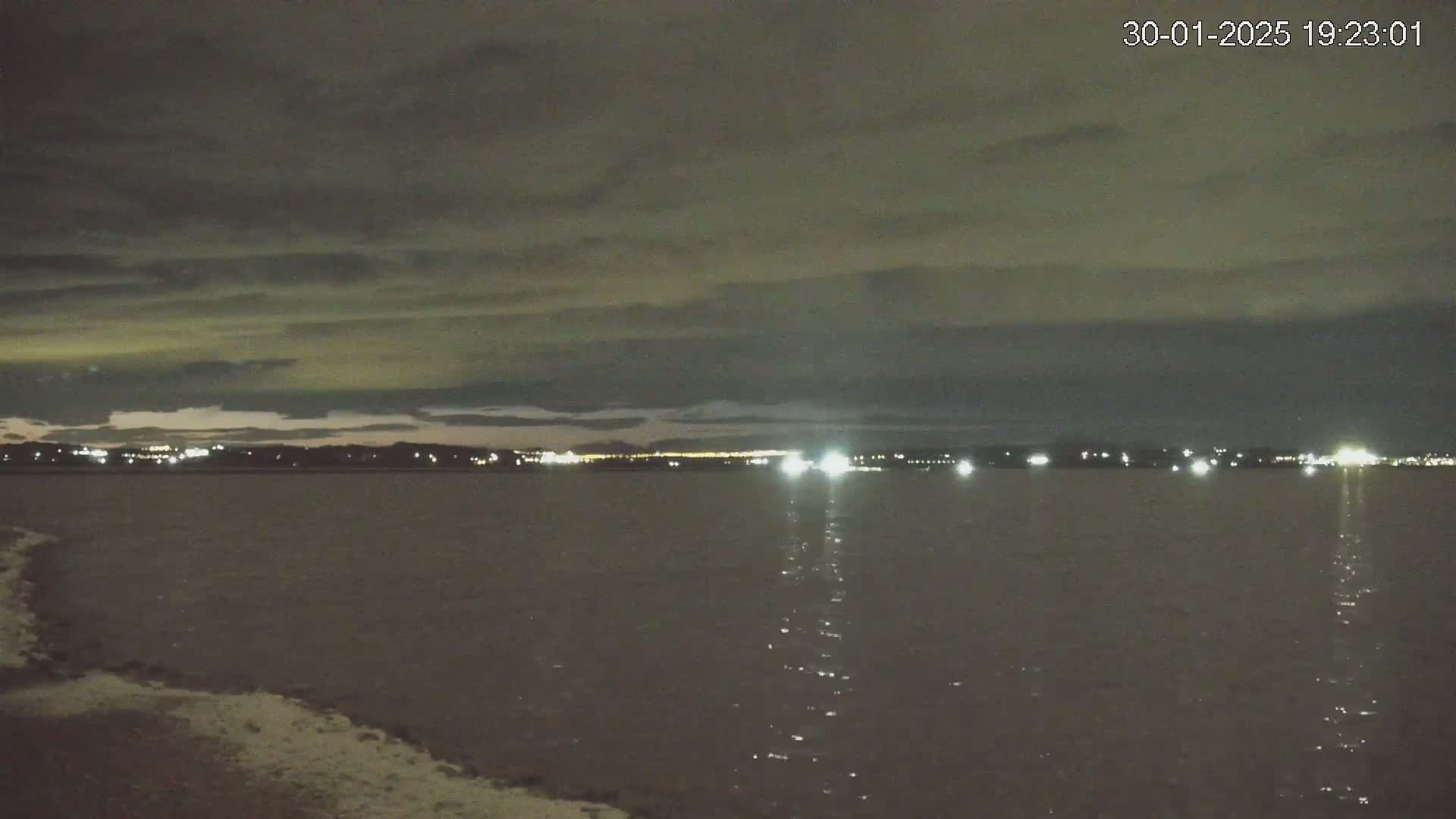 A nighttime view of a calm body of water with a distant, faintly lit city skyline under a cloudy sky.