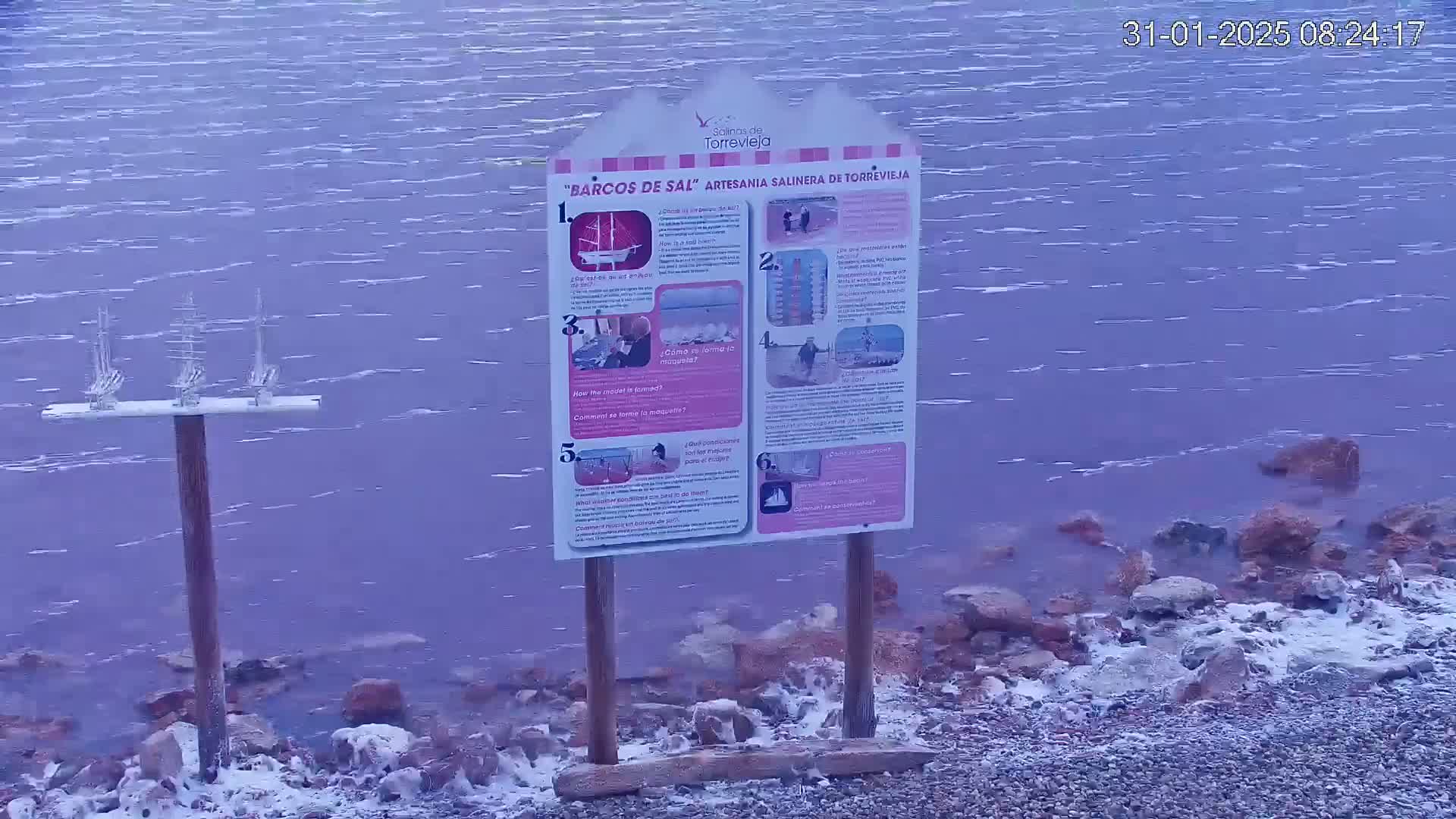 A body of purplish water with small waves laps against a rocky shore dusted with frost, near an informational sign and a display of small, white, ship-like objects.