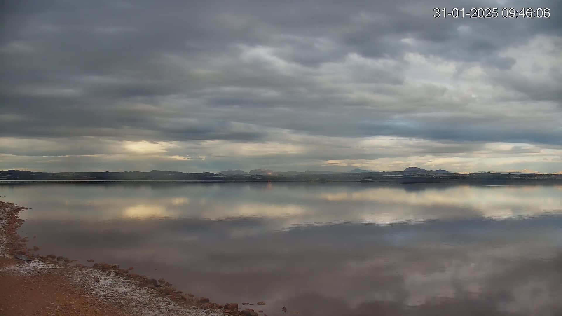 A calm body of water reflects a cloudy sky and distant mountains under overcast conditions.