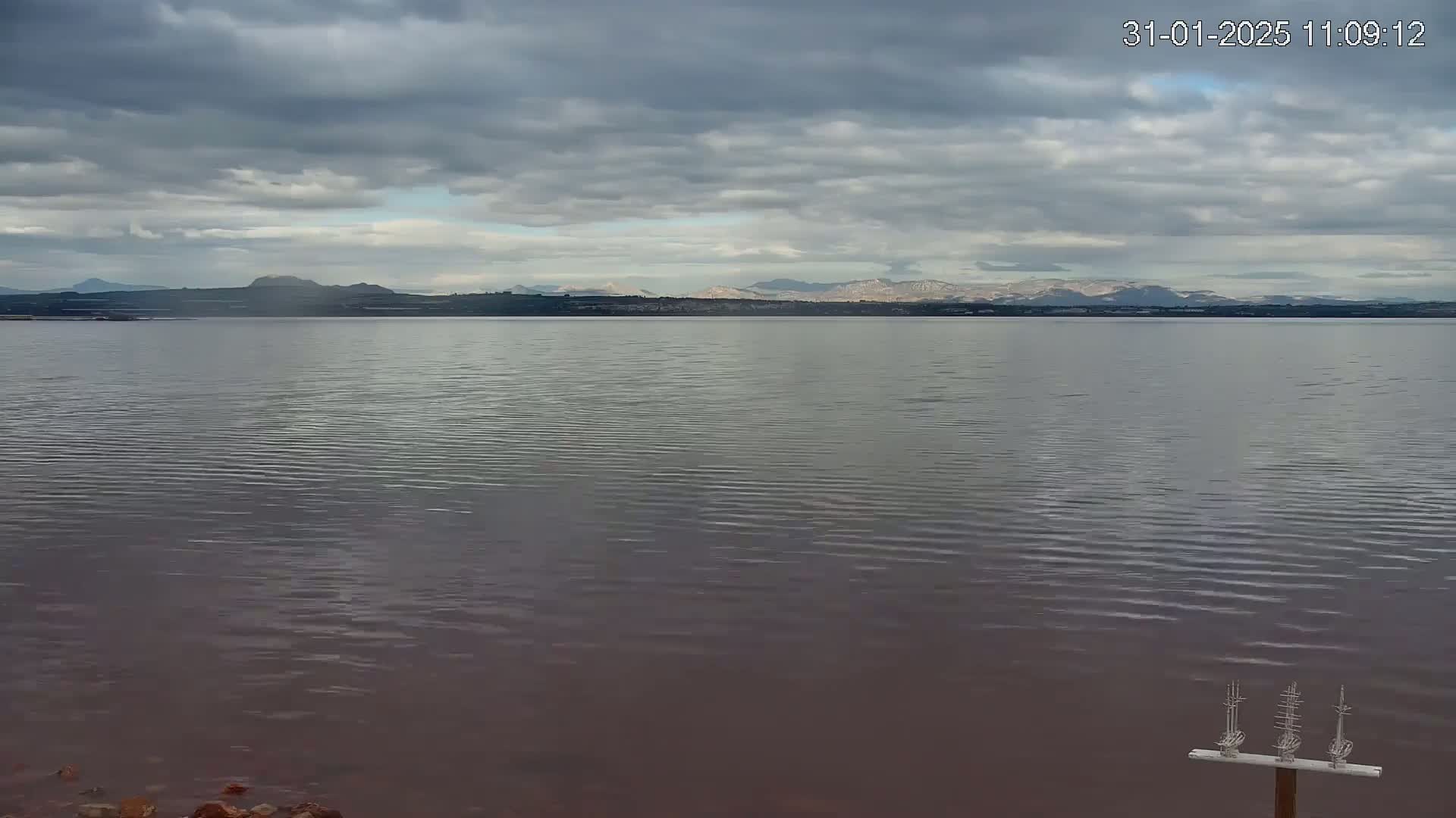 A calm body of water with a slightly reddish tint stretches towards a distant mountain range under a cloudy sky.