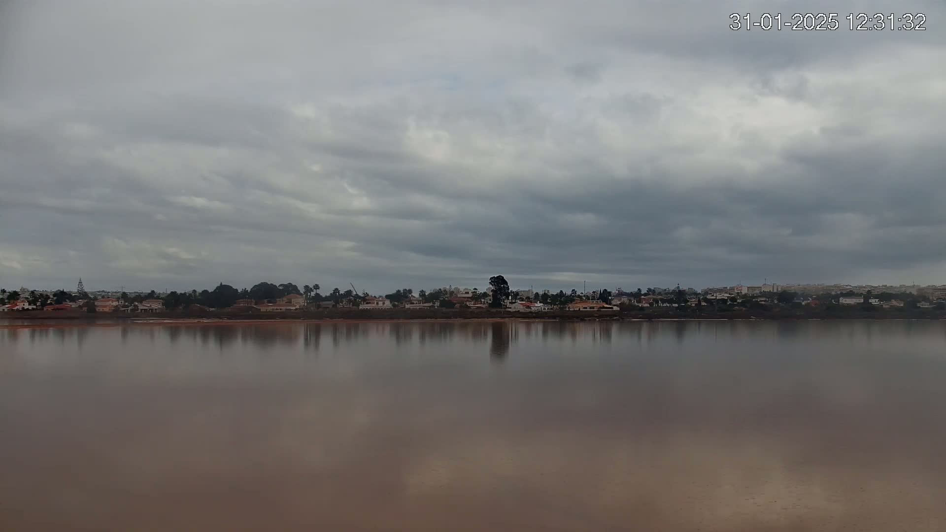 A calm body of water reflects a distant town under a cloudy sky.