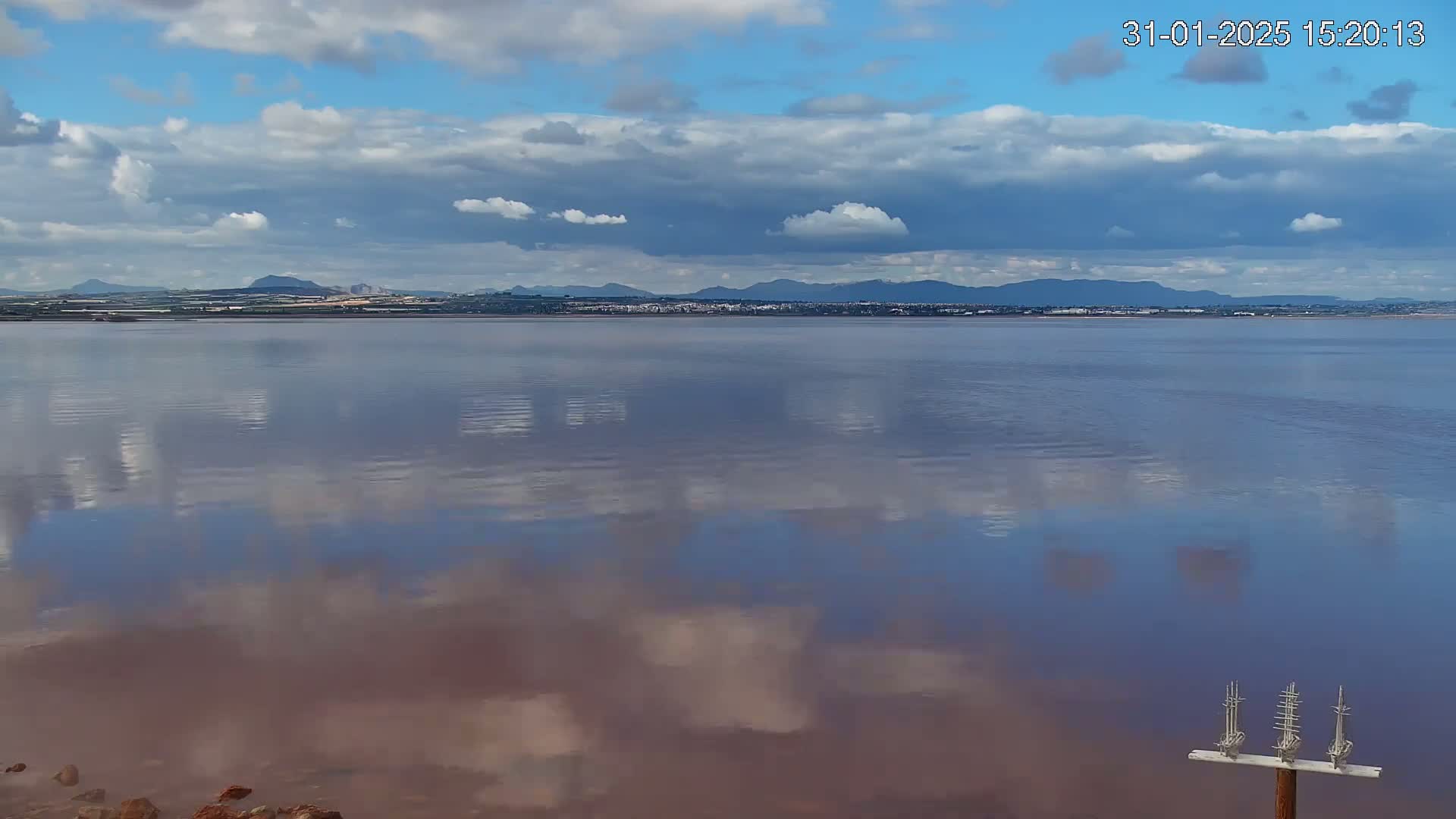 A calm body of reddish-pink water reflects a partly cloudy sky and distant mountains under a mostly clear sky.