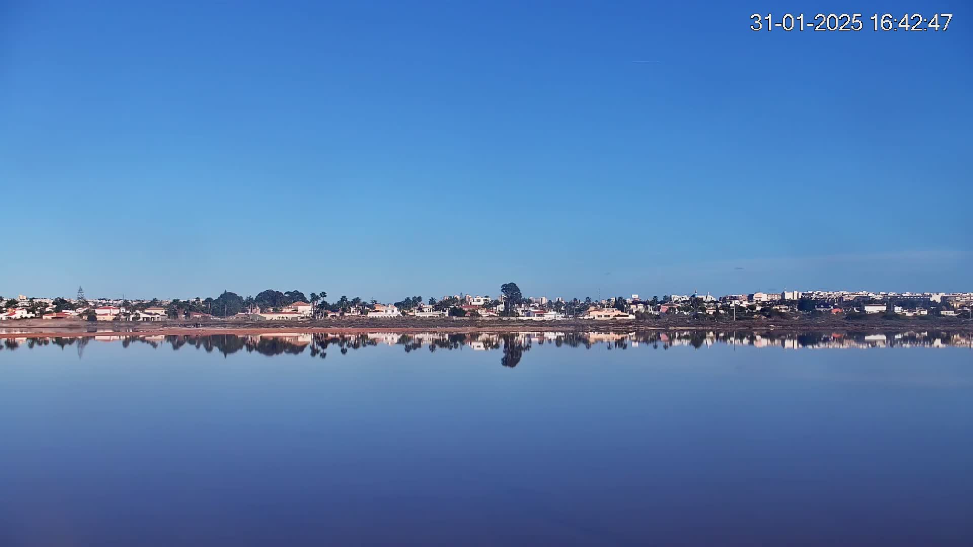 A calm, clear blue sky reflects on a still body of water, mirroring a distant shoreline of houses and trees.