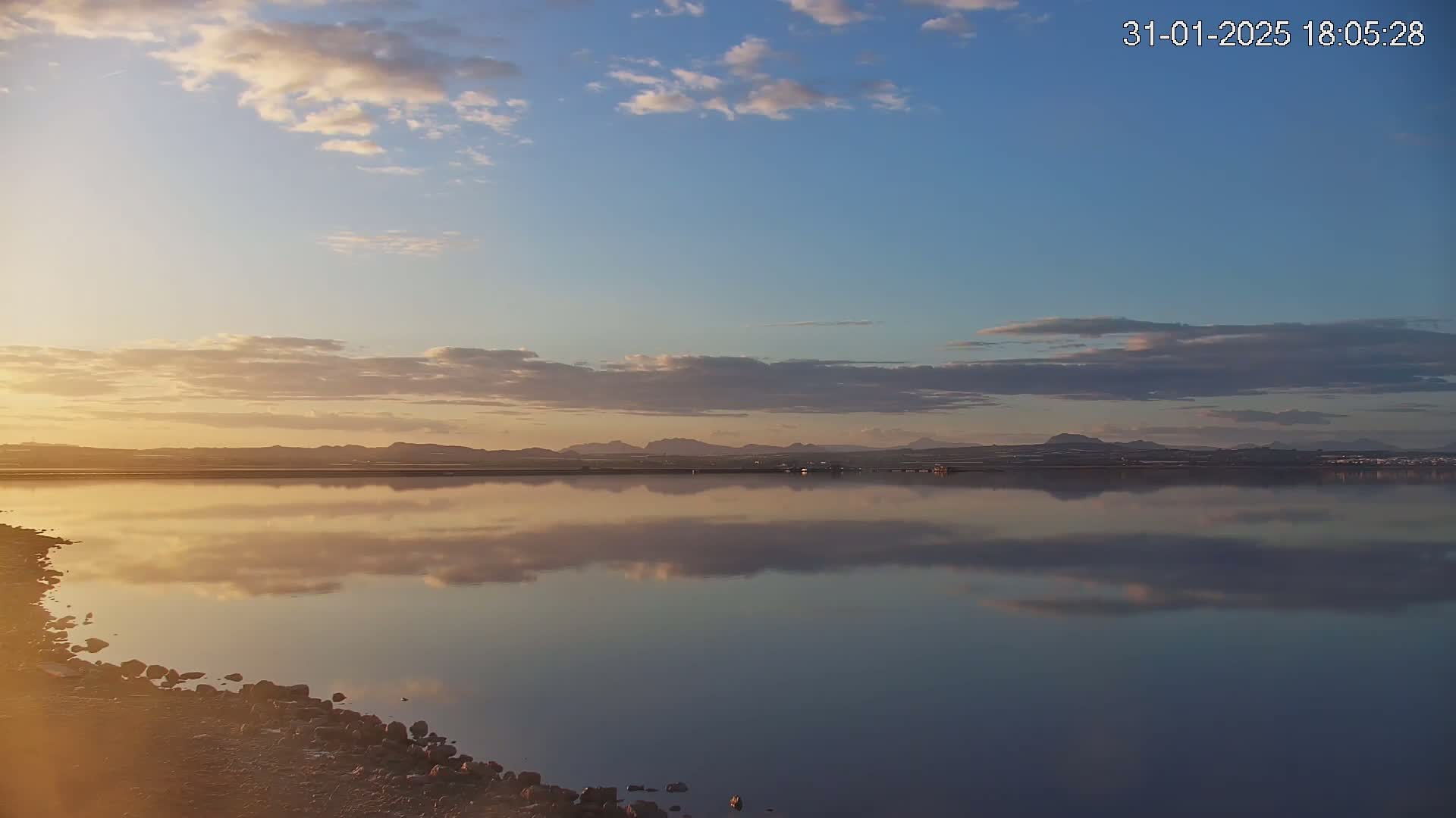 A calm body of water reflects a sunset sky and distant mountains under a partly cloudy sky.
