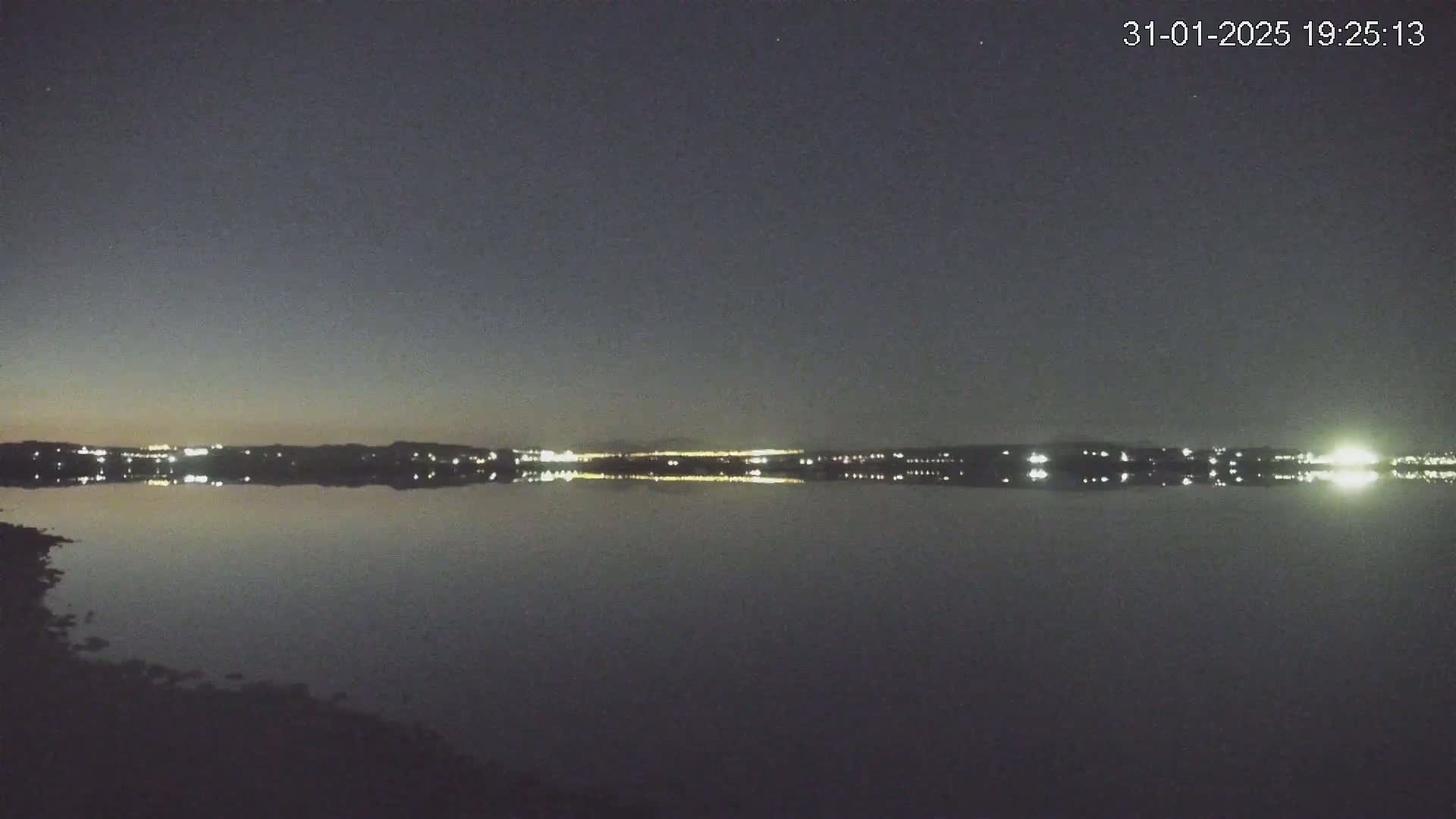A calm body of water reflects the lights of a distant city under a dark, twilight sky.