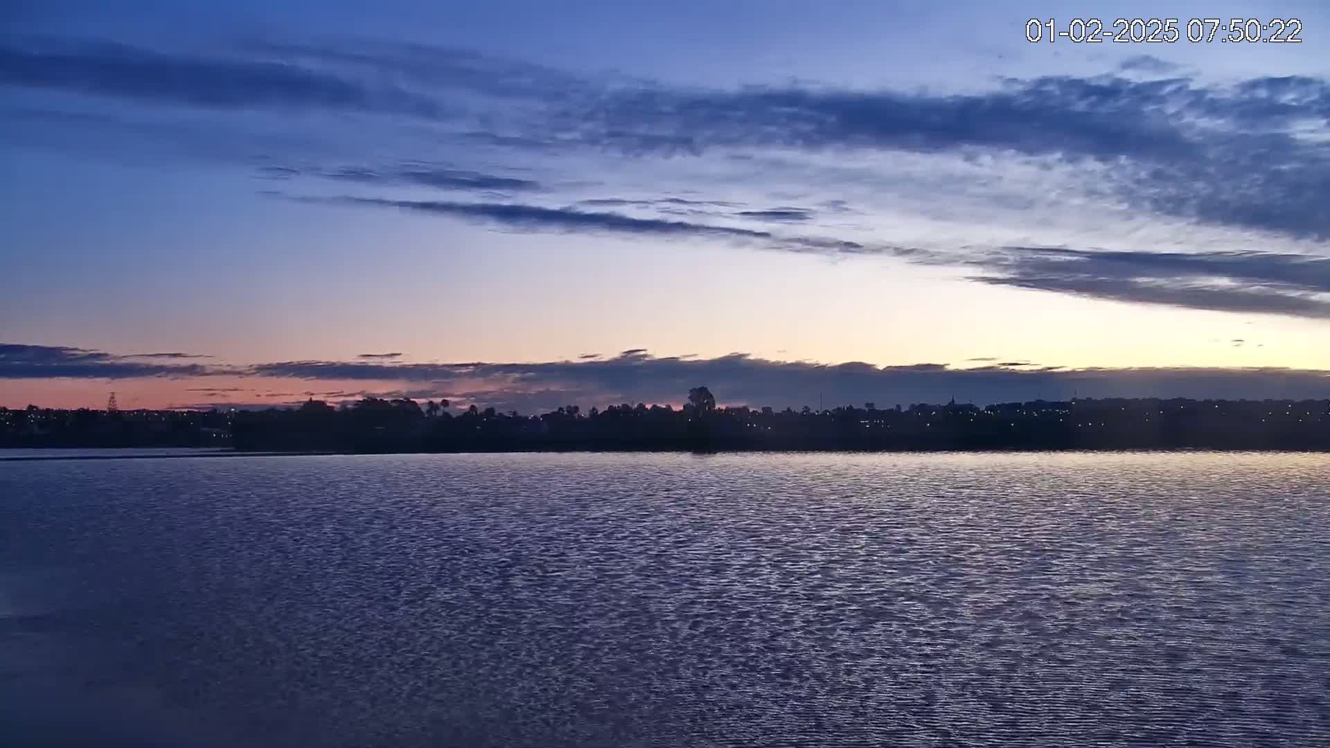 A calm body of water reflects the dusky colors of a sunrise sky under a mostly cloudy sky.