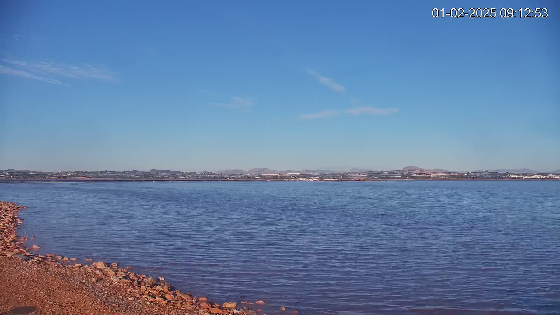 A calm body of water with a rocky shore stretches towards a distant mountainous horizon under a clear blue sky.