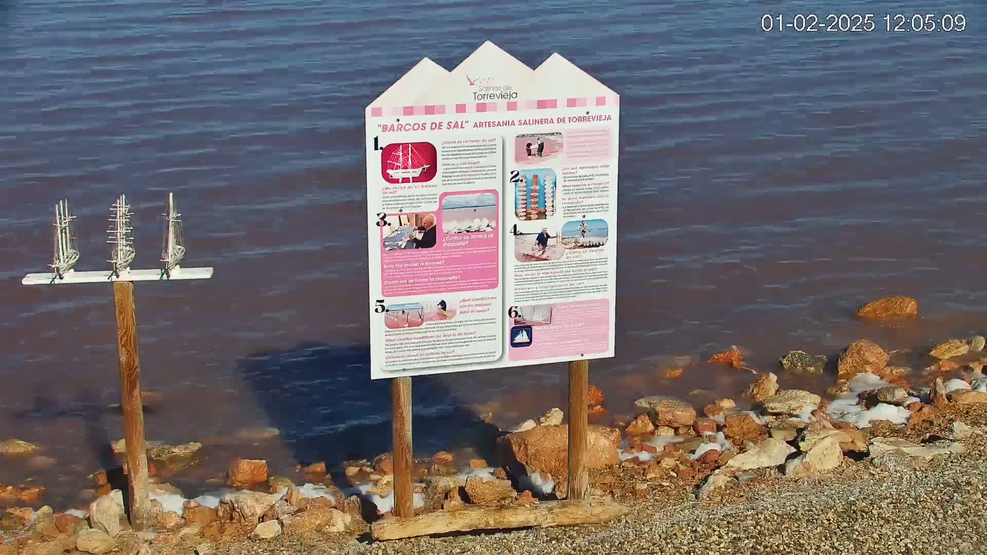A wooden sign with illustrations and descriptions stands near a pinkish-red body of water with small waves lapping a rocky shore, and three model sailboats are mounted on a post behind it.
