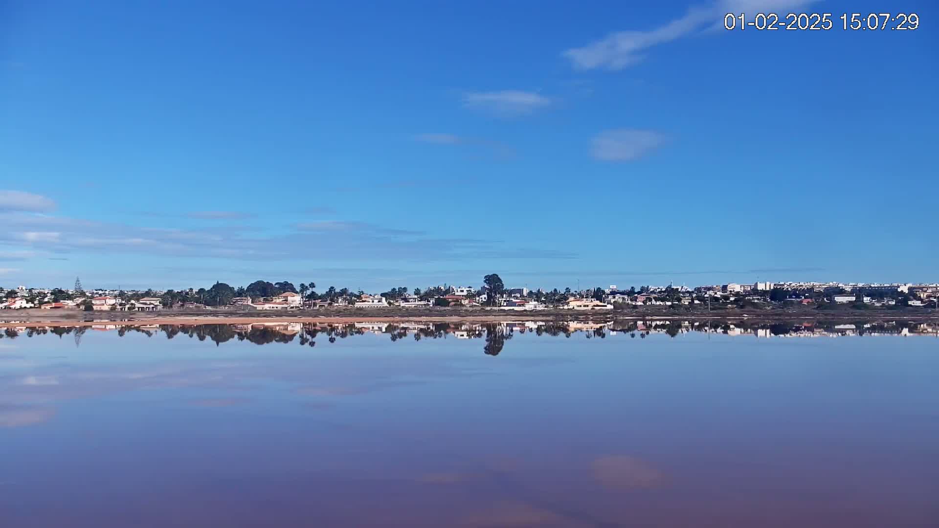 A calm, pink-hued body of water reflects a clear blue sky and a line of houses and vegetation on the opposite shore under a mostly clear sky.