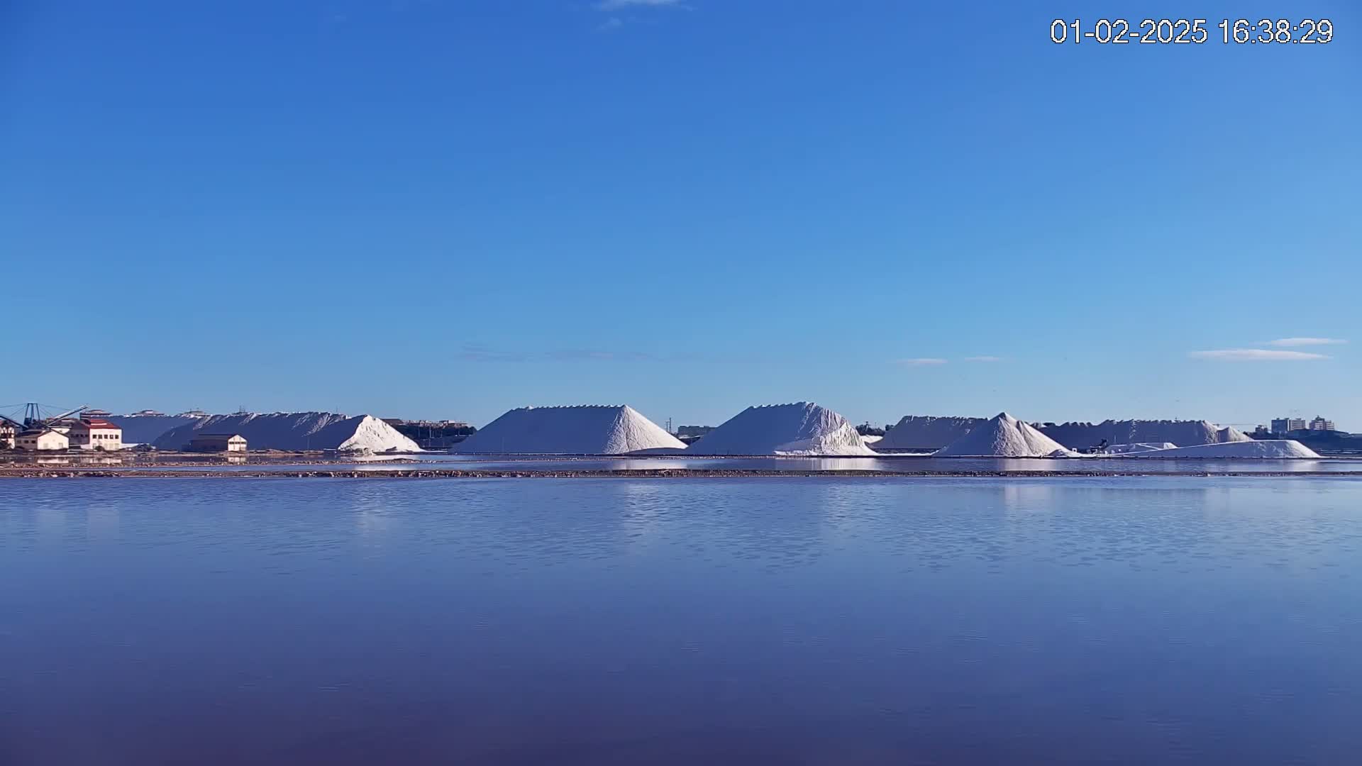 Under a clear blue sky, several large piles of white salt are situated next to a calm, purplish-blue body of water.