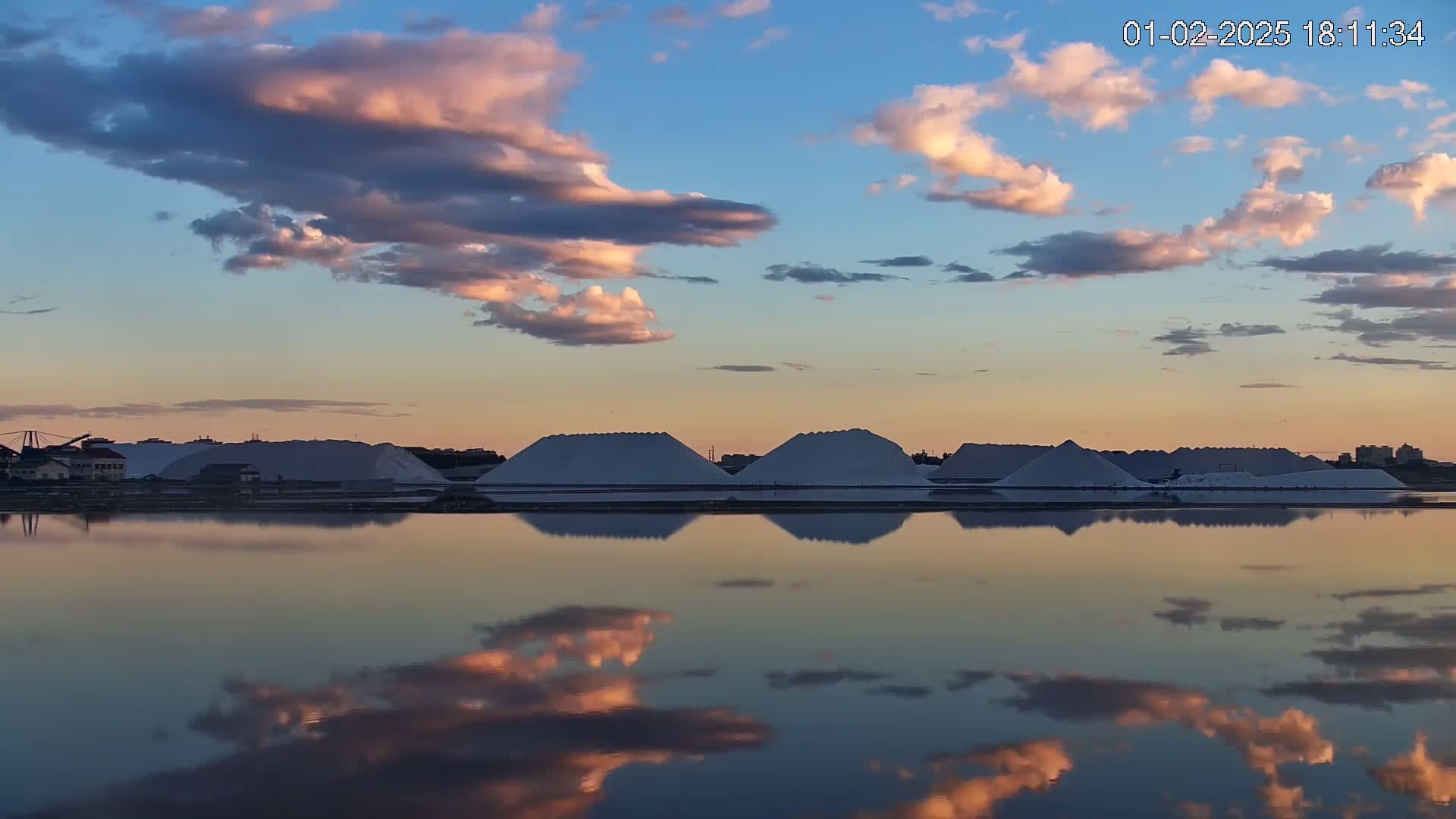 Large piles of white salt are reflected in a calm body of water under a sunset sky filled with pink and blue clouds.