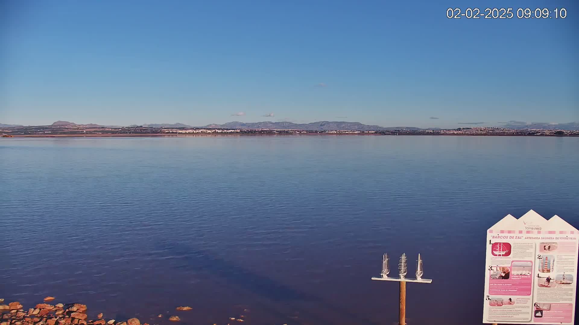A calm body of water stretches toward a distant town nestled against a mountain range under a clear blue sky.