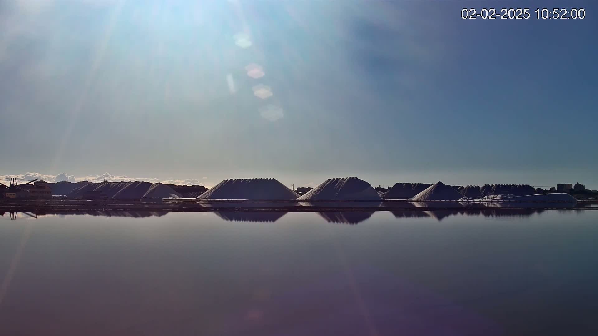 A calm body of water reflects a row of large, conical piles of salt under a bright, sunny sky.