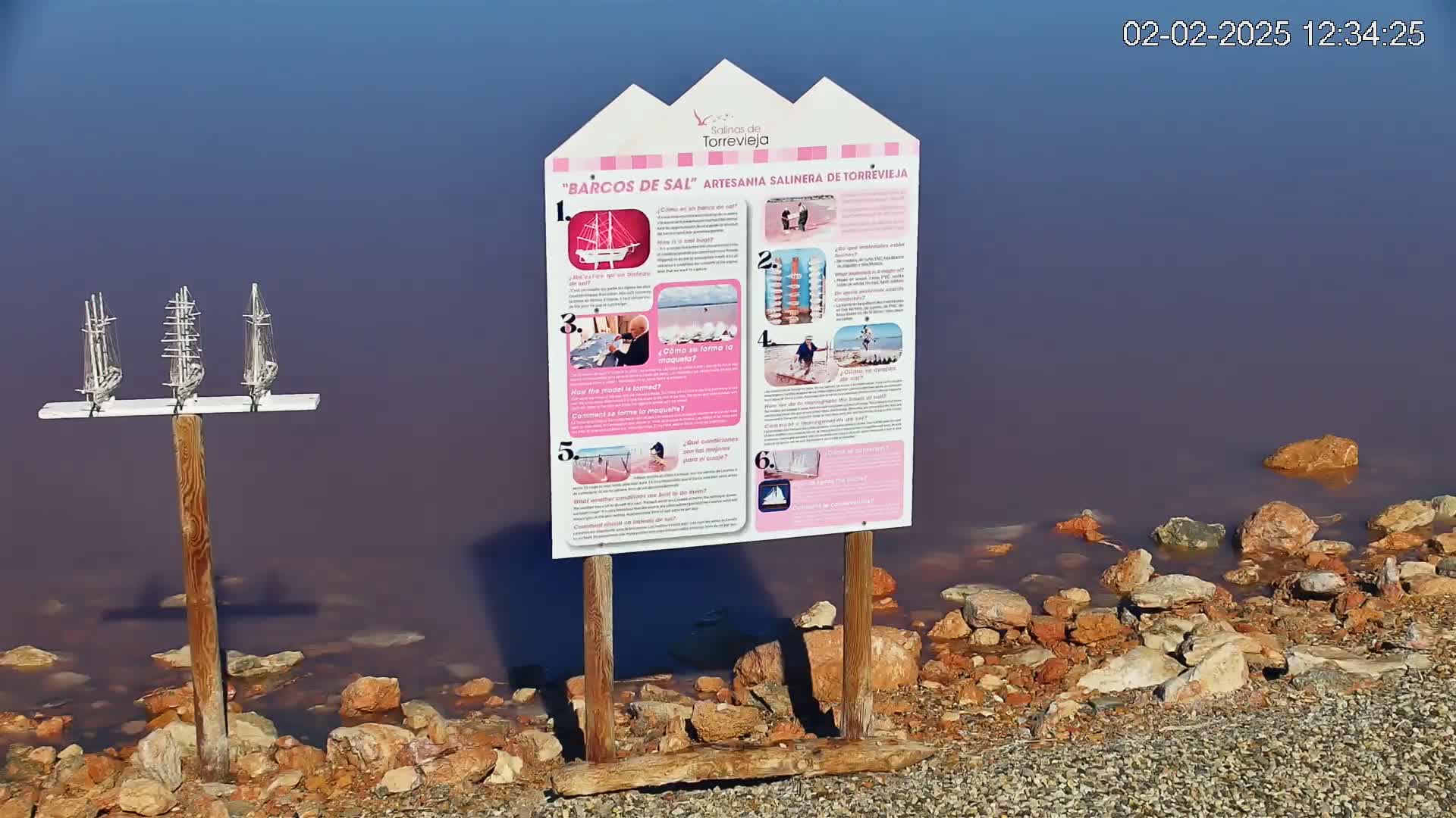 A sign with pictures stands near a pinkish body of water, rocks, and a display of miniature sailboats on a wooden post.
